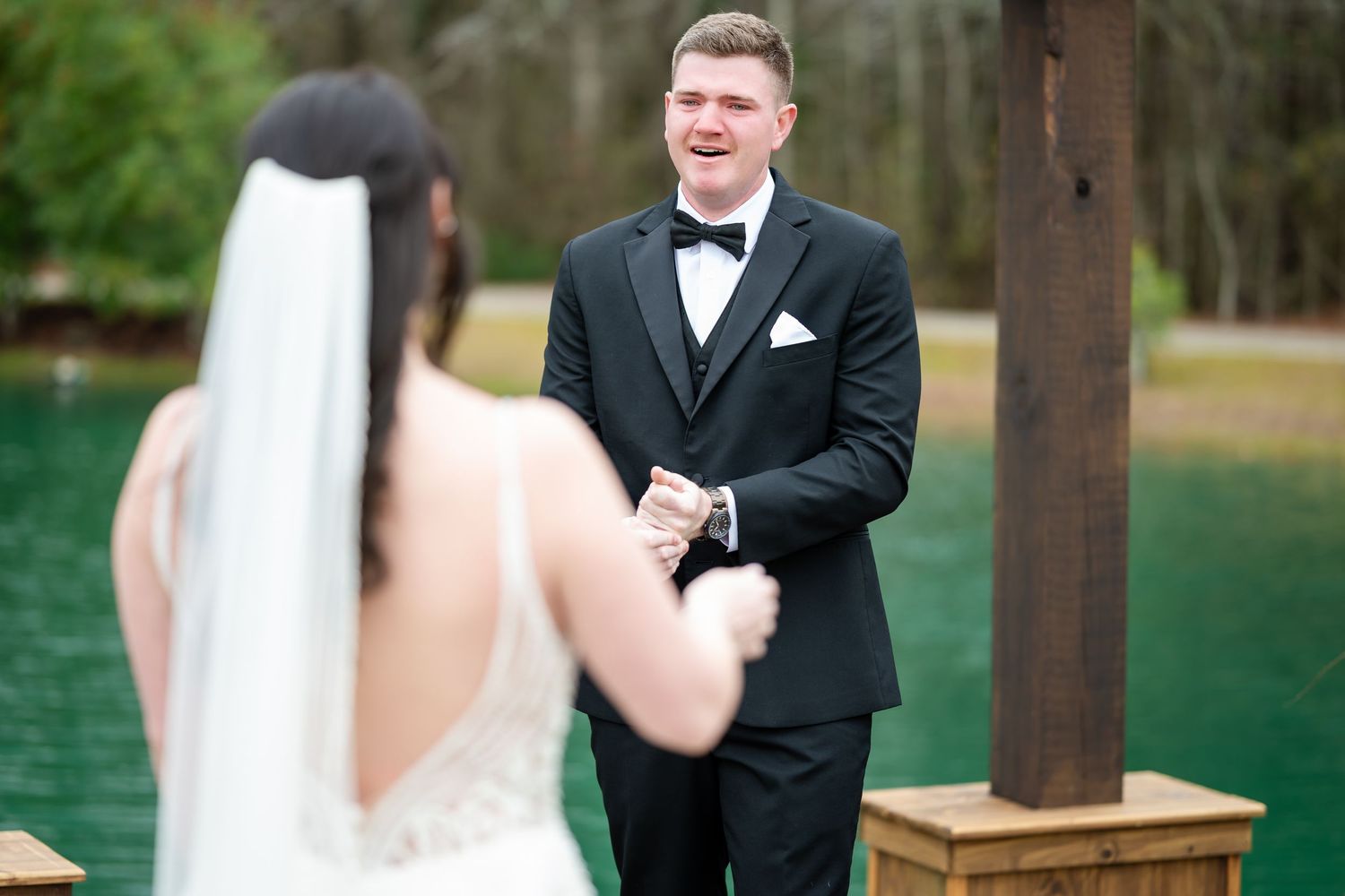 Emotional groom smiling at bride during outdoor first look by turquoise lake.