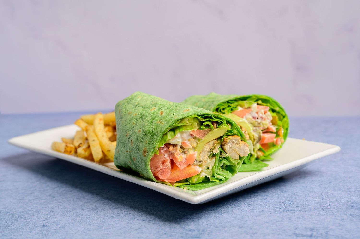 Green spinach wrap sandwich cut in half, filled with fresh vegetables and protein, served with golden french fries on a white plate.