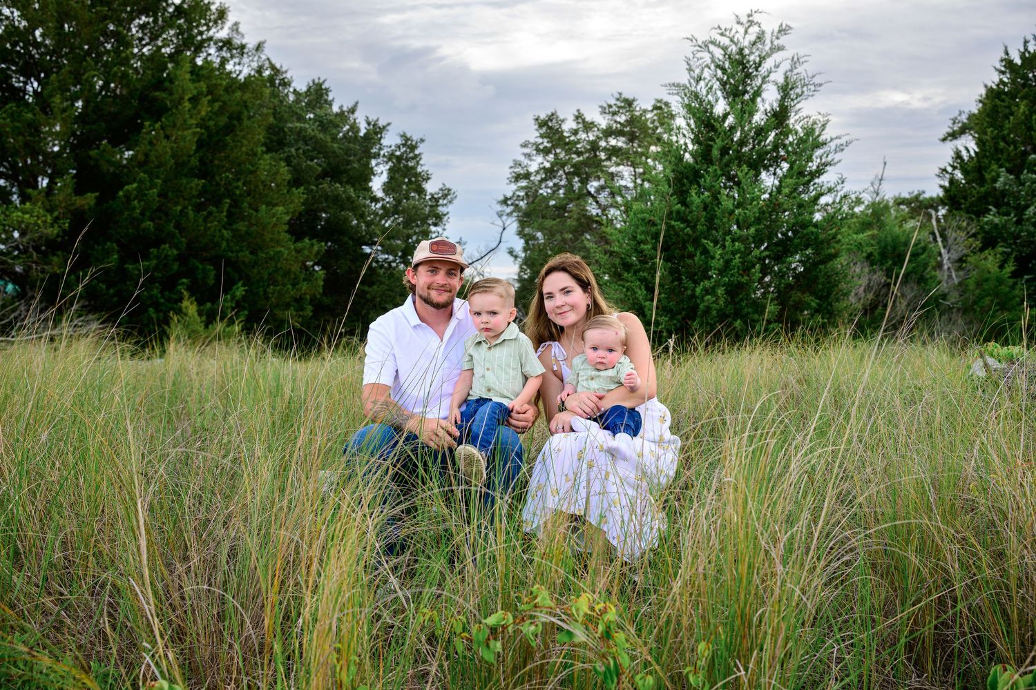 Happy family with two young children poses together in tall green grass field for outdoor portrait.