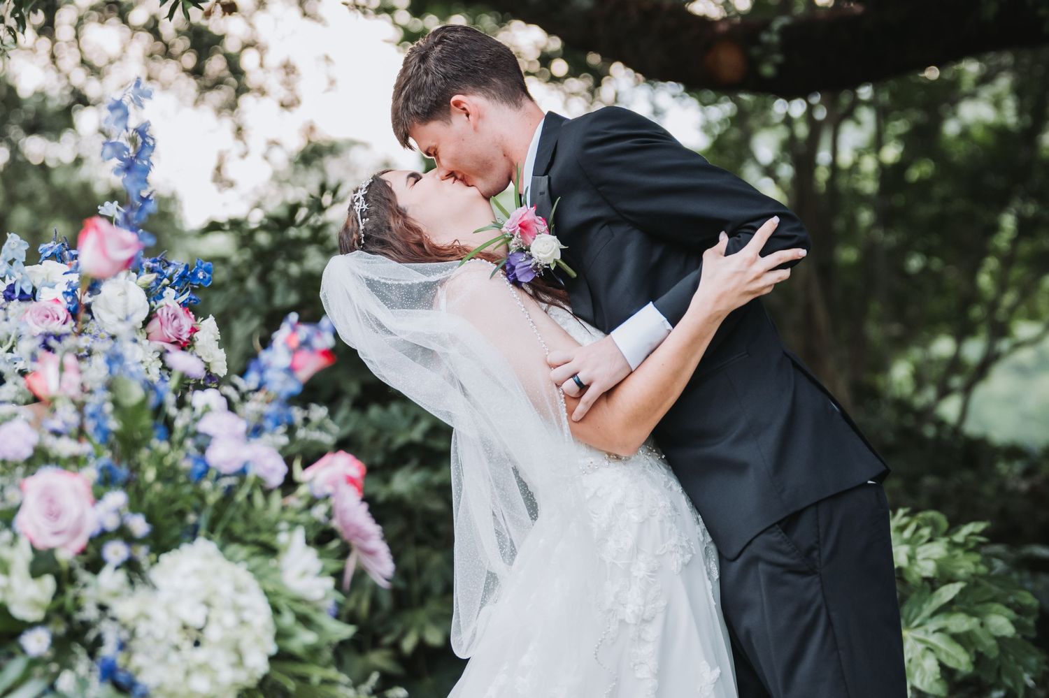 Bride and groom share first kiss as newlyweds surrounded by colorful floral wedding arch.