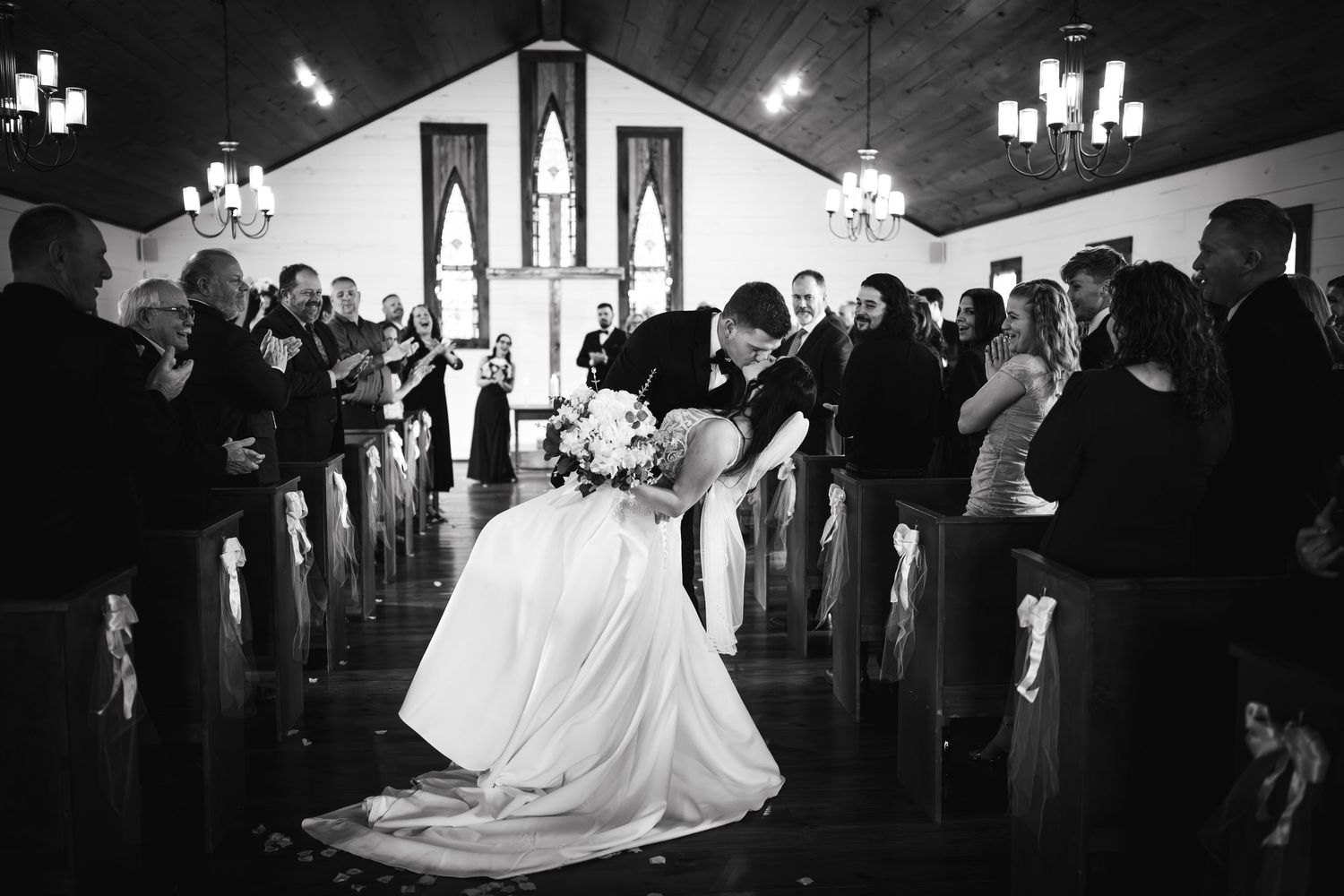Black and white photo of newlyweds sharing dramatic dip kiss in rustic chapel aisle.