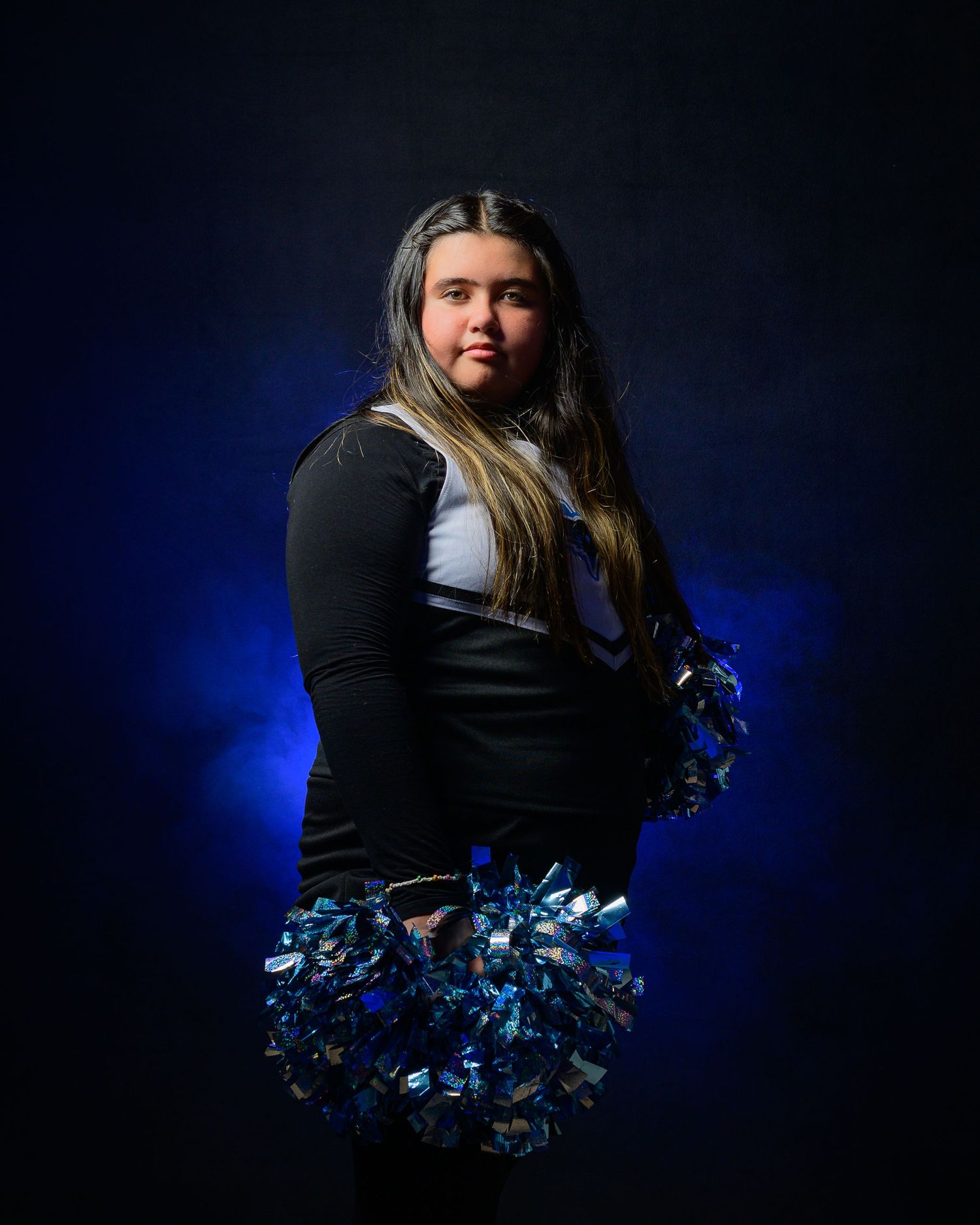 Young cheerleader in black and white uniform holds blue metallic pom-poms against dark blue backdrop.