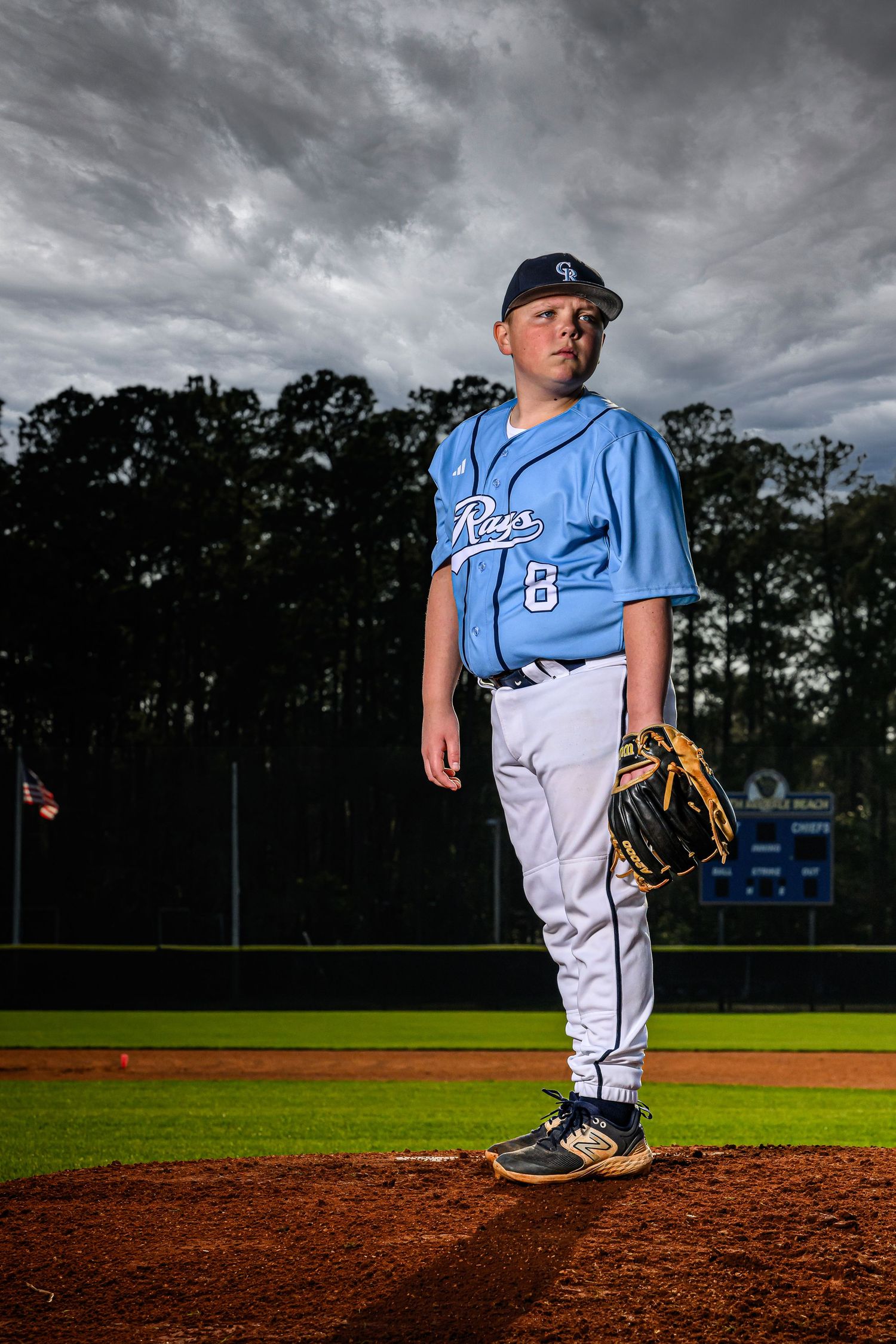 Young baseball pitcher in blue Rays uniform stands on mound with glove under dramatic cloudy sky.