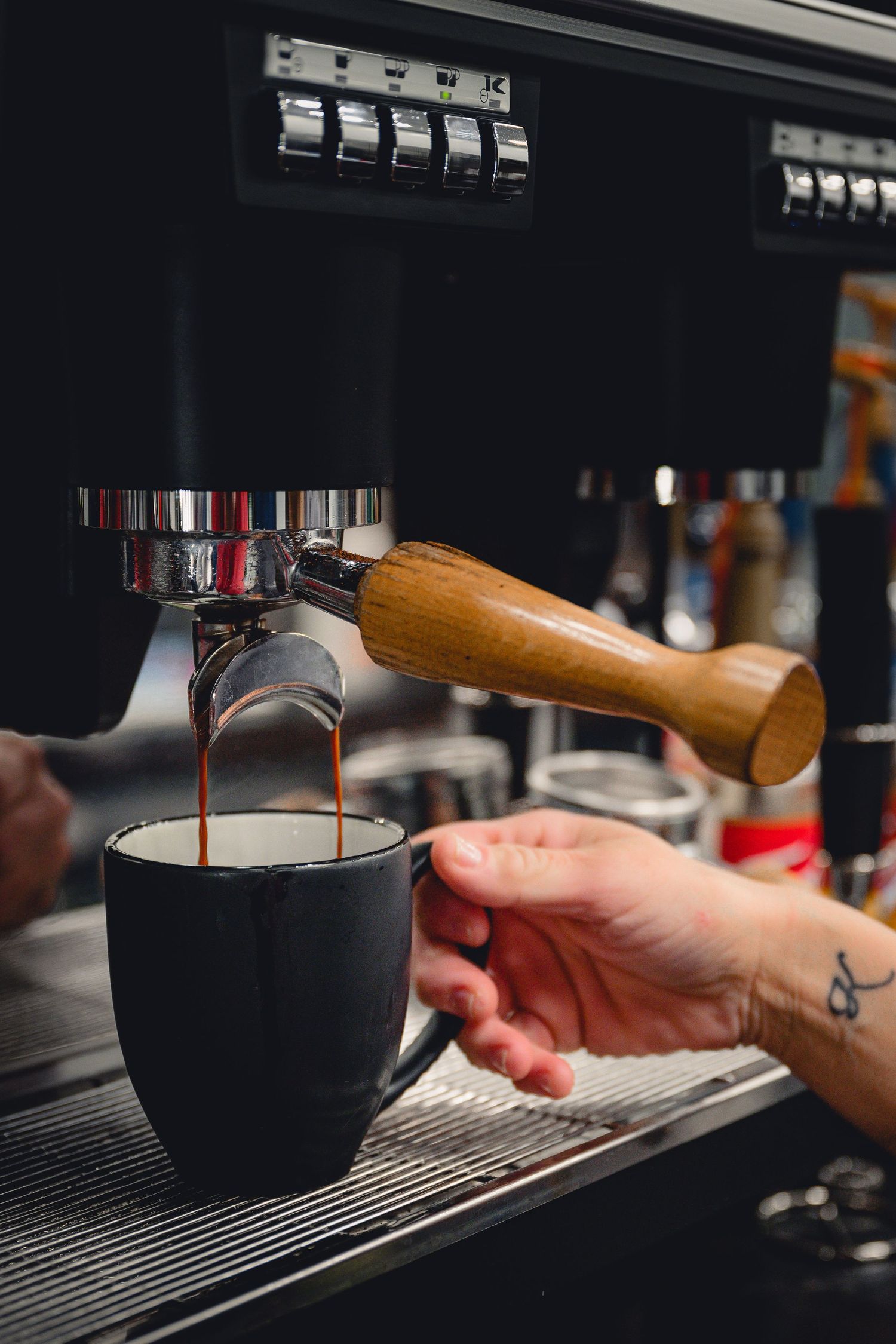 Barista pulling espresso shot with wooden-handled portafilter into black mug.