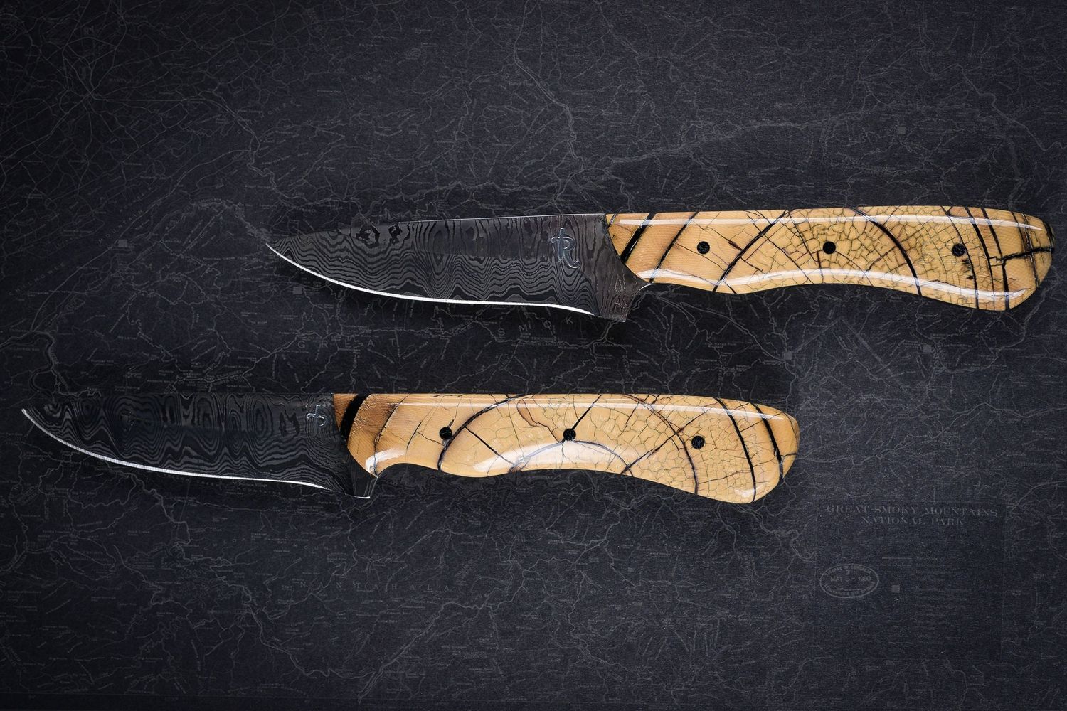 Two handmade fixed blade knives with Damascus steel blades and natural wood handles on dark stone.