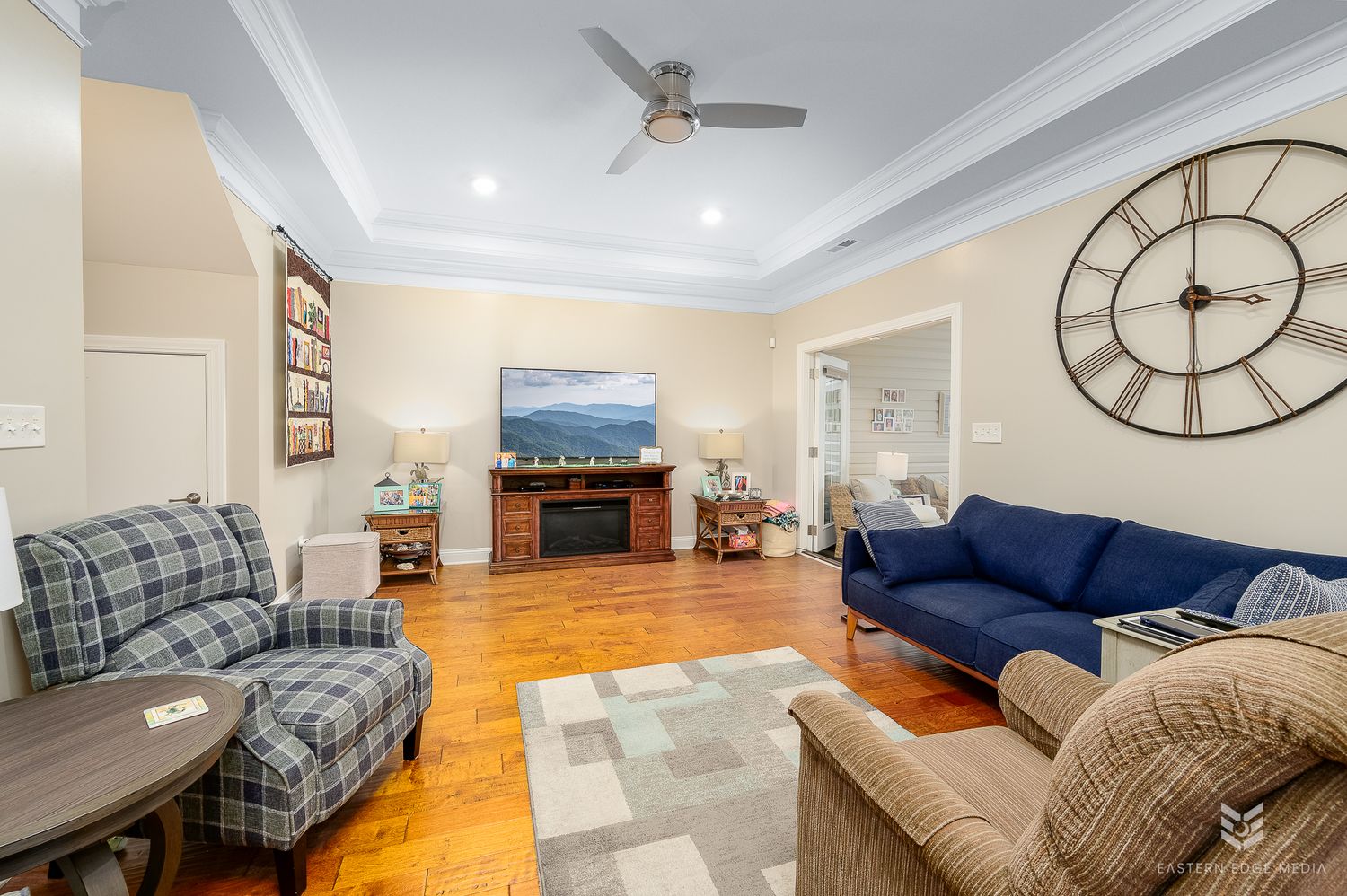 Spacious living room featuring tray ceiling, ceiling fan, fireplace, blue sofa, and plaid armchairs on hardwood floors.