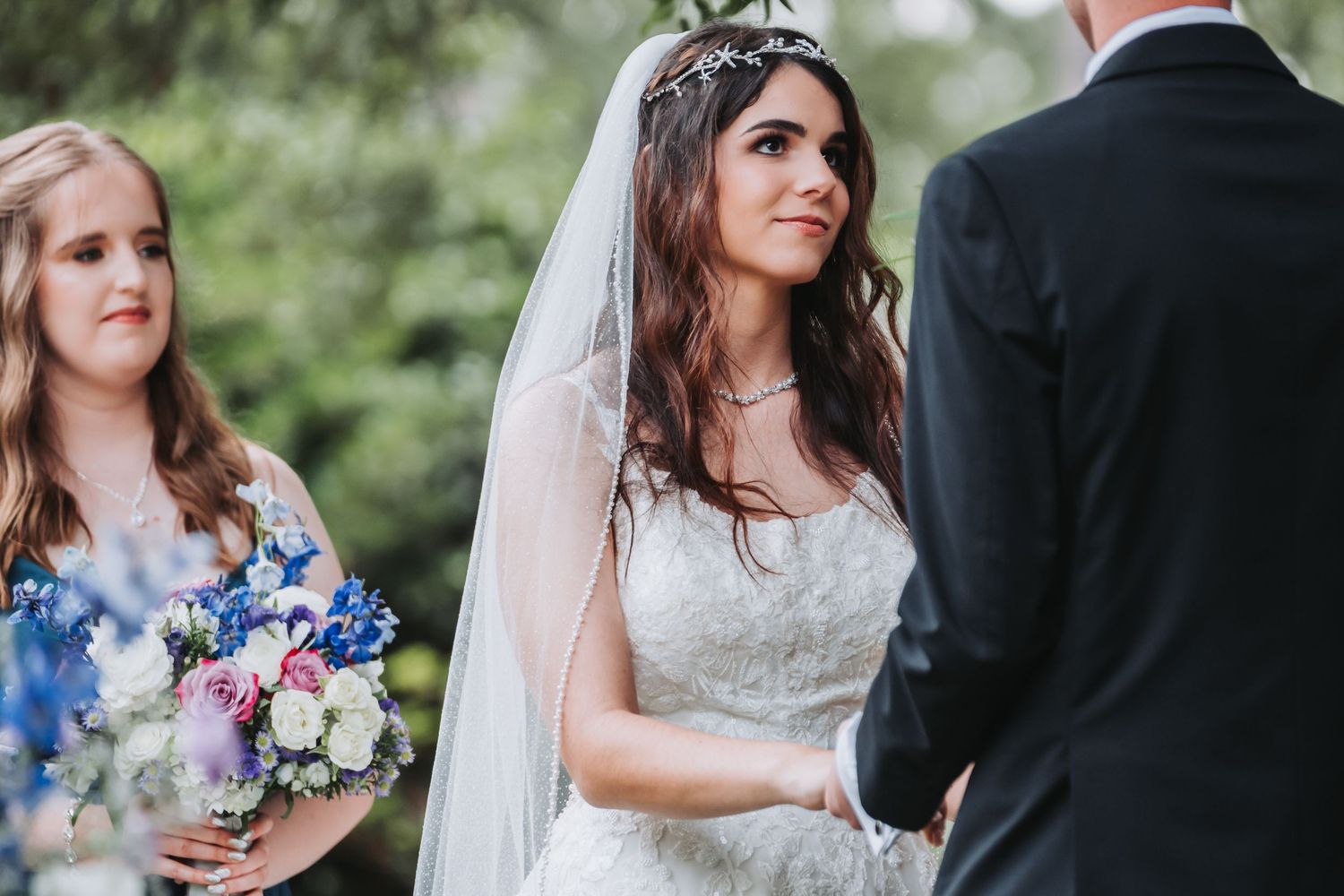 Bride gazes lovingly at groom during outdoor wedding ceremony, bridesmaid holding bouquet behind her.