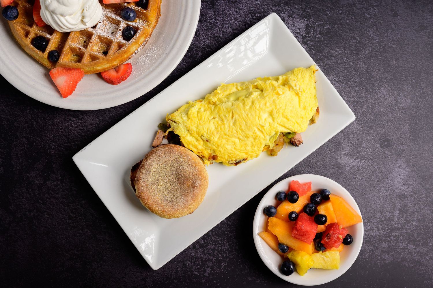 Fluffy yellow omelette served with an English muffin and fresh fruit bowl alongside a waffle with berries and whipped cream.