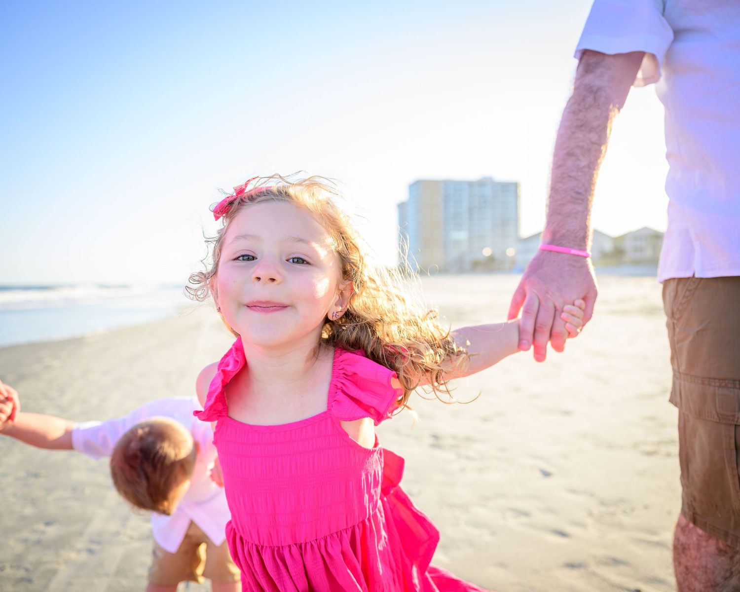 Happy young girl in pink dress holding fathers hand on sunny beach.