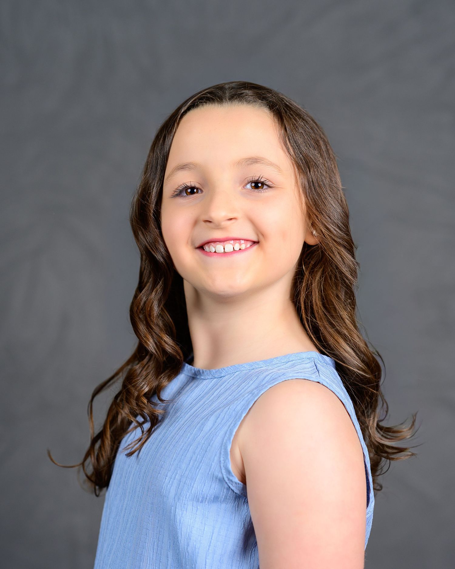 Smiling young girl with curly brown hair poses in blue sleeveless top against gray backdrop.