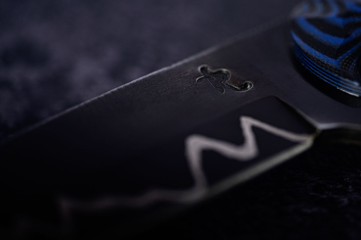 Close-up macro shot of engraved black Damascus steel knife blade with blue handle detail.