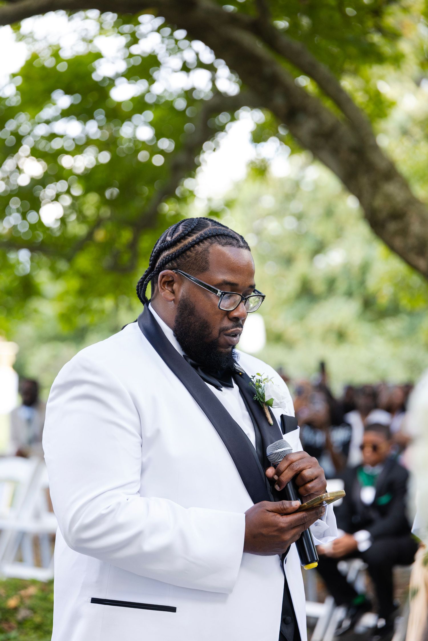 A groom with braided hair and glasses wears a white tuxedo jacket and reads from a phone into a microphone at an outdoor wedding.