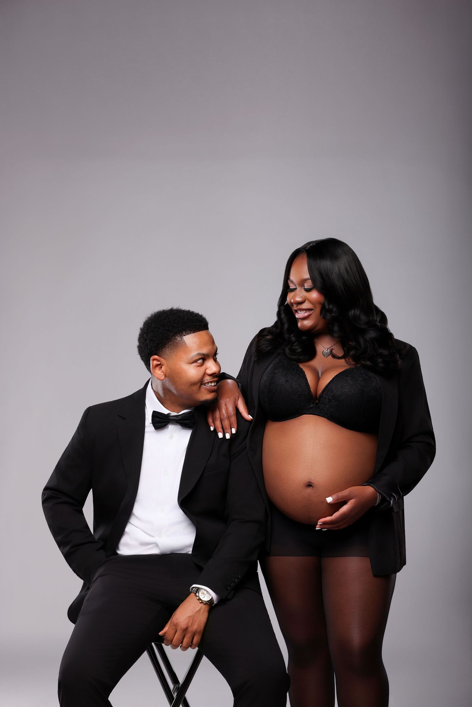 Smiling expectant couple in a studio maternity portrait, the man in a black tuxedo and bow tie seated beside his standing pregnant partner.
