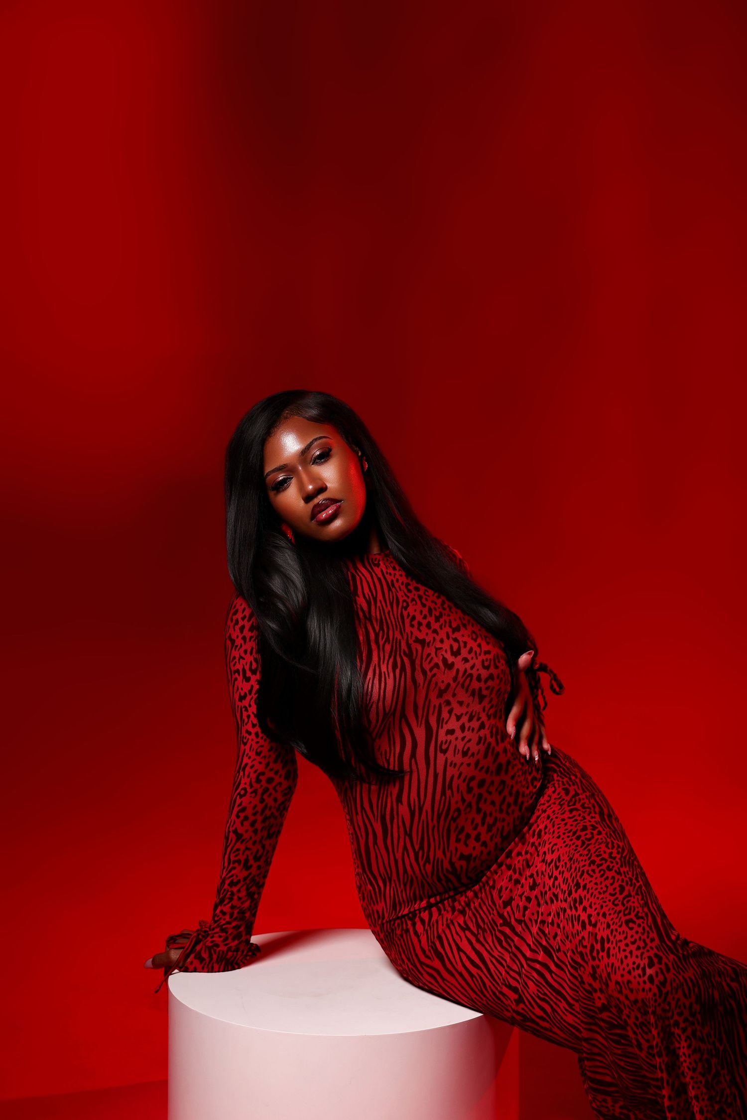 A woman in a red animal print gown poses on a white pedestal against a bold red studio backdrop in a dramatic fashion portrait.