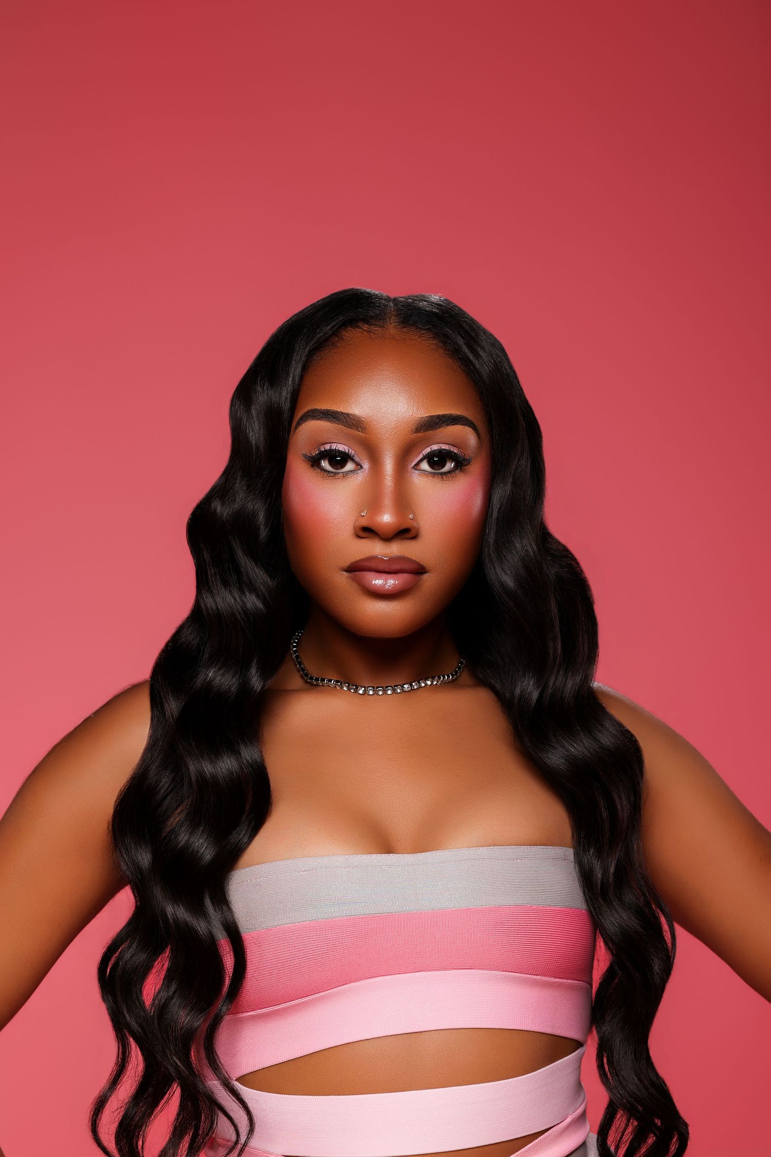 Woman with long wavy black hair poses against a pink backdrop wearing a pink and gray striped bandeau top and a choker necklace.