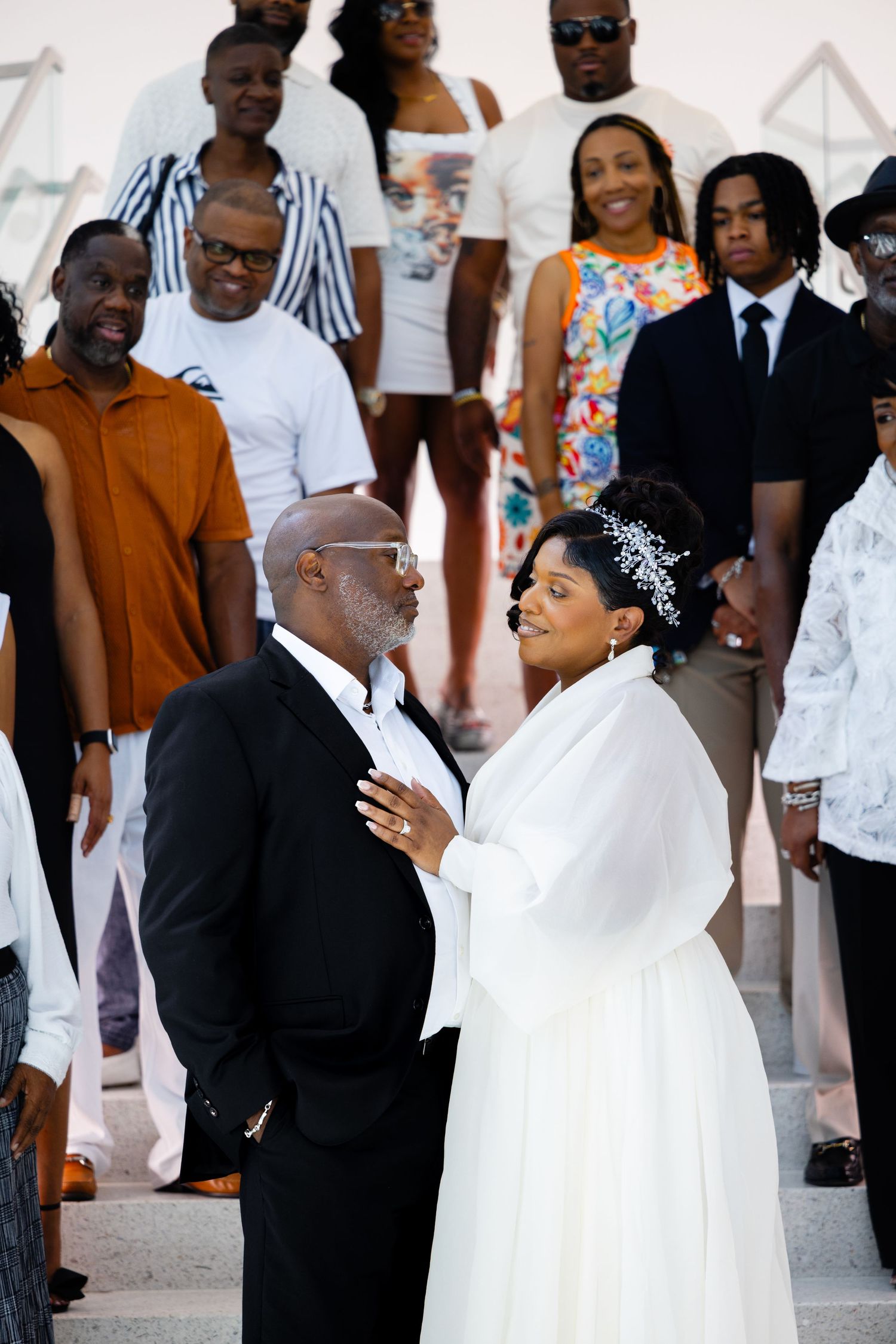 Bride in a white gown and floral hairpiece rests her hand on a groom's chest as smiling wedding guests look on from the steps behind them.
