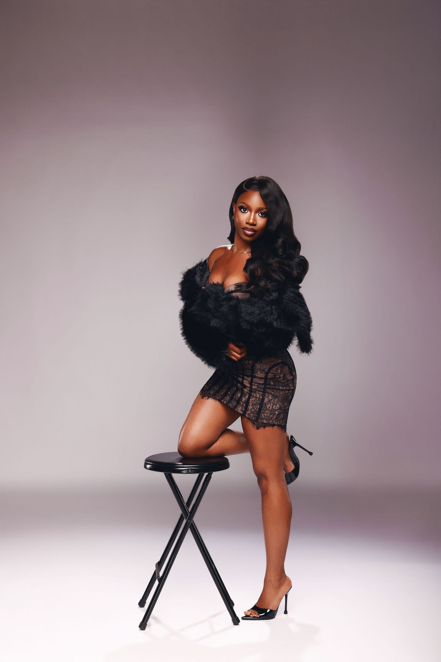 A woman in a black lace dress with a fluffy feather wrap poses beside a black stool in heels against a soft purple studio backdrop.