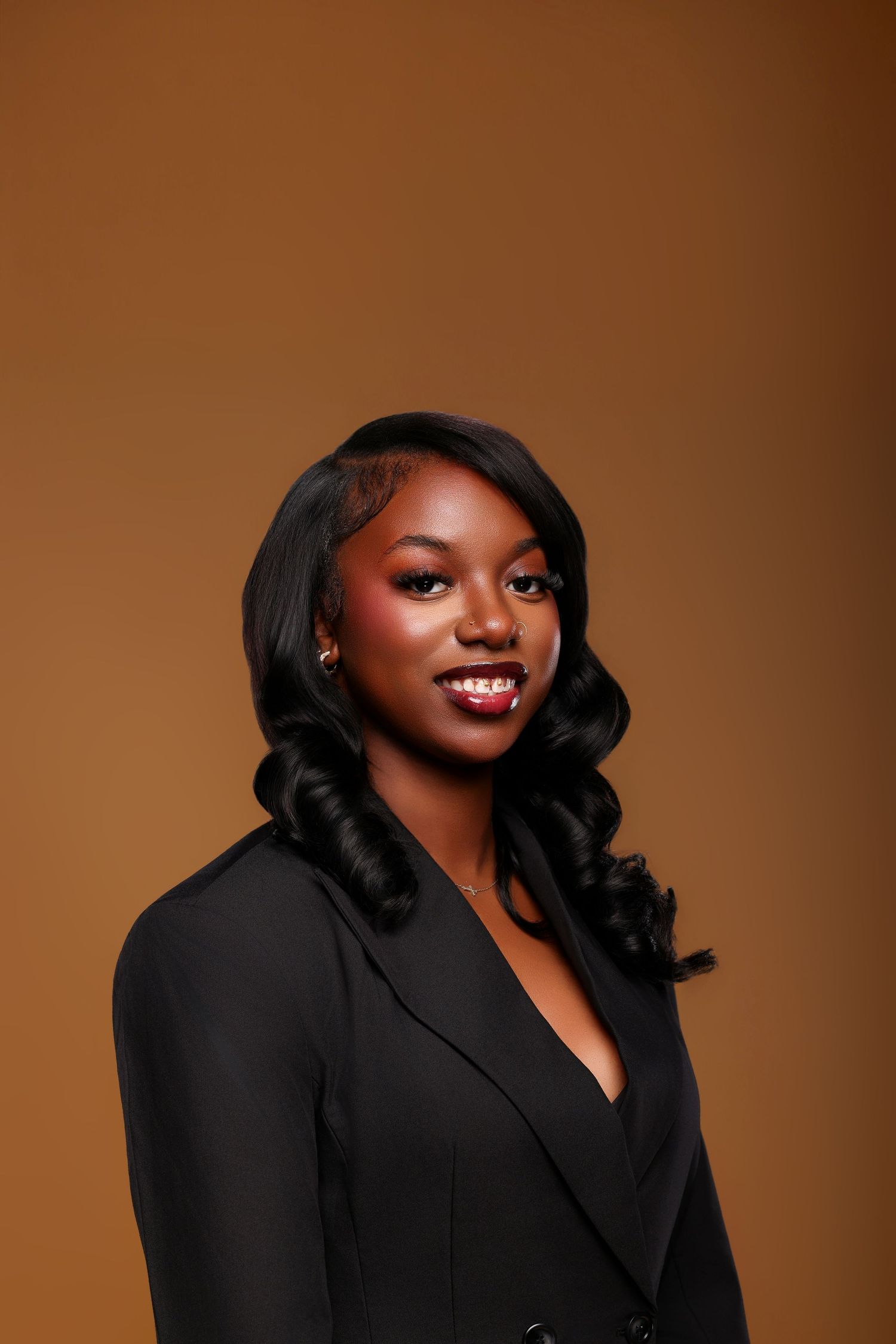 Smiling woman with long black curled hair wearing a black blazer poses for a professional portrait against a warm golden-brown backdrop.