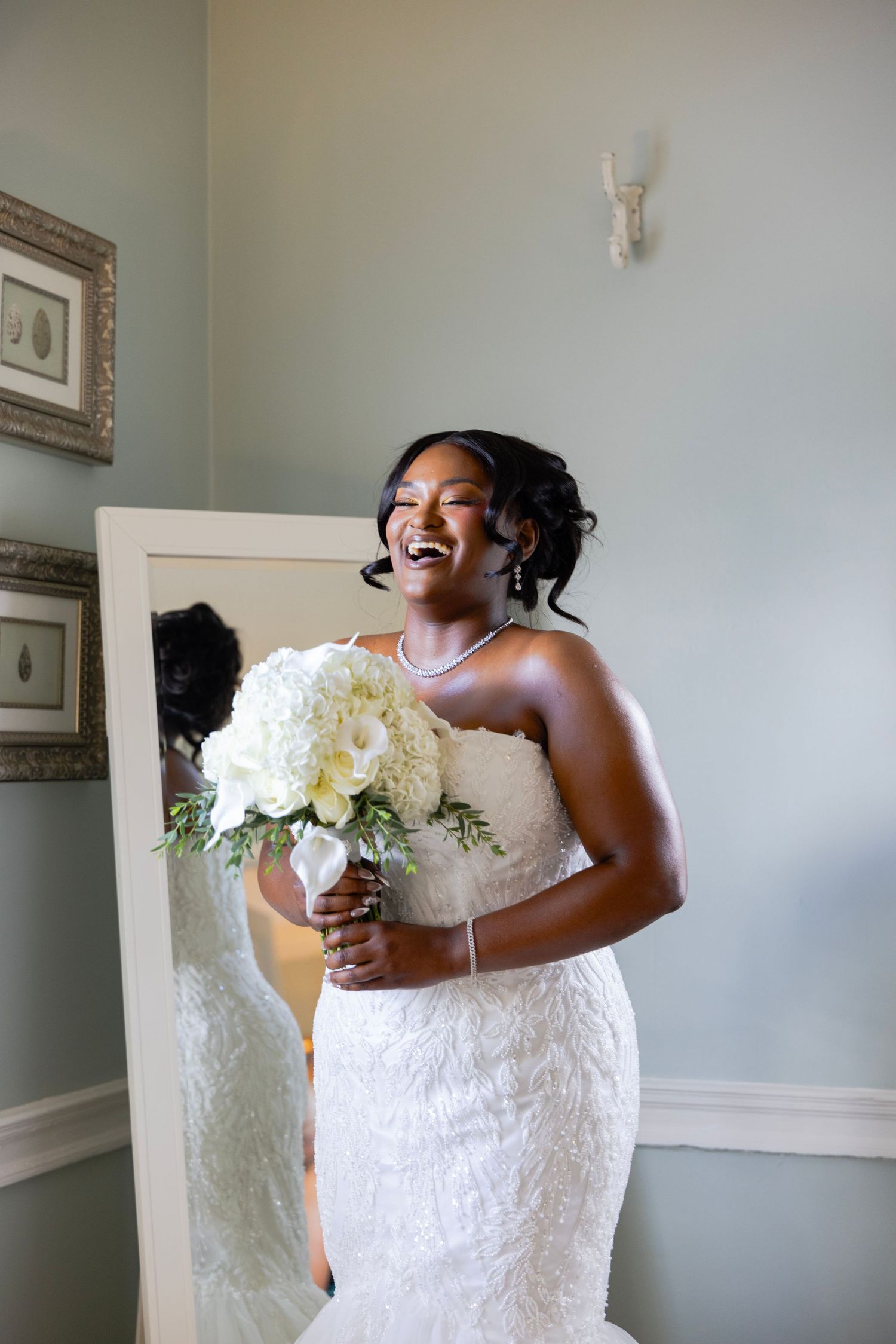 A smiling bride in a strapless white lace gown laughs while holding a white floral bouquet beside a tall mirror.