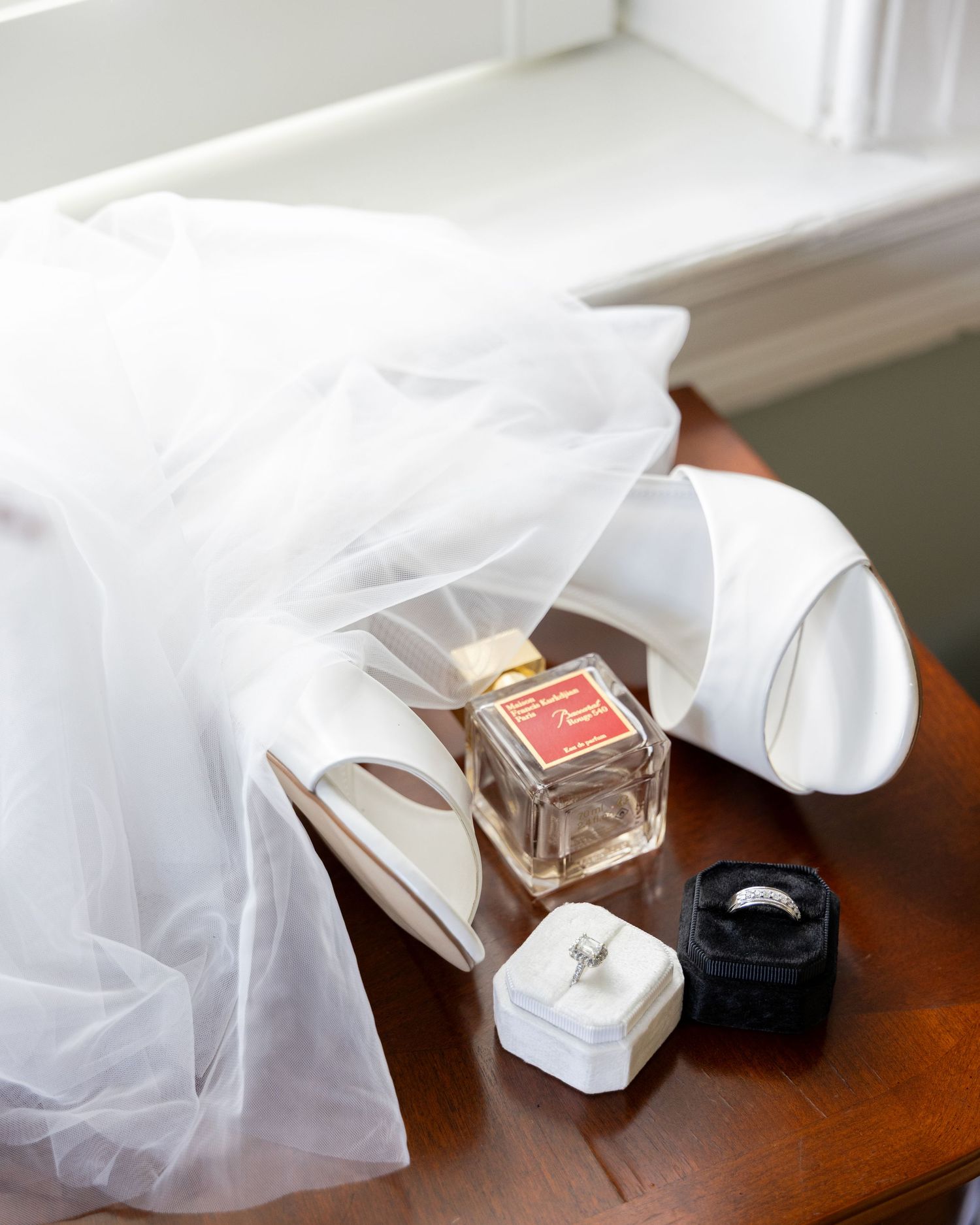 Bridal details featuring white heels draped in tulle veil, a perfume bottle, and two wedding ring boxes on a wooden table by a window.