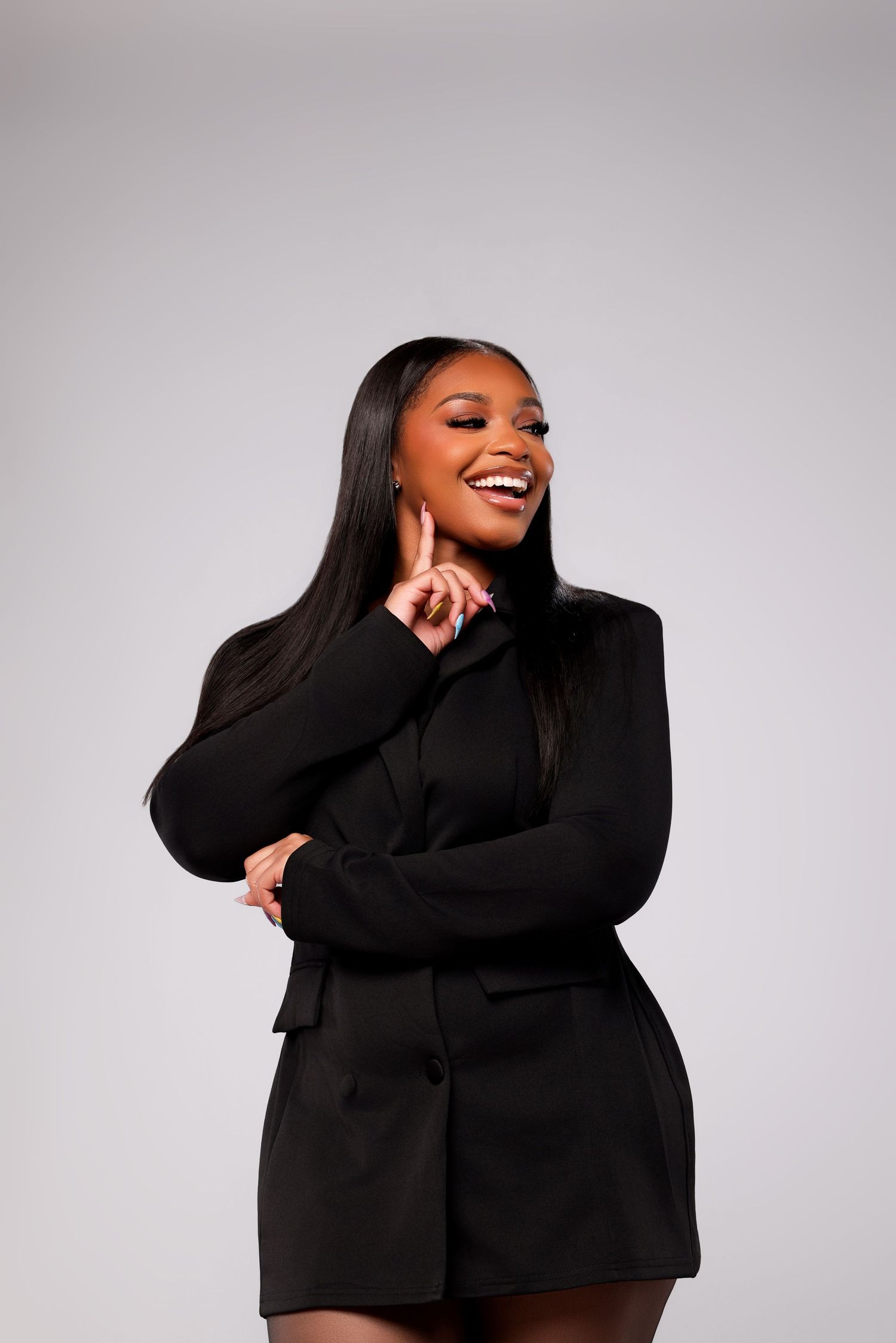 A smiling woman with long straight hair wears a black blazer dress and strikes a playful pose against a light gray studio backdrop.