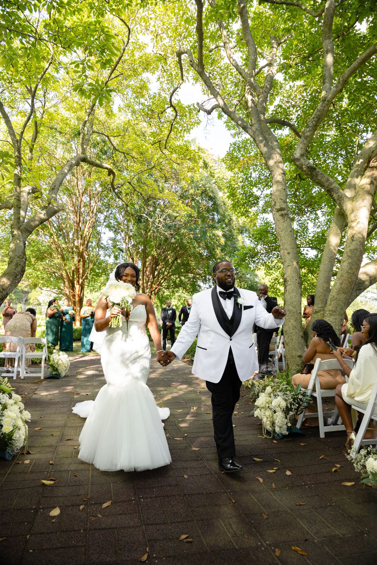 A bride and groom hold hands and walk down a tree-lined brick aisle as seated wedding guests look on after the ceremony.