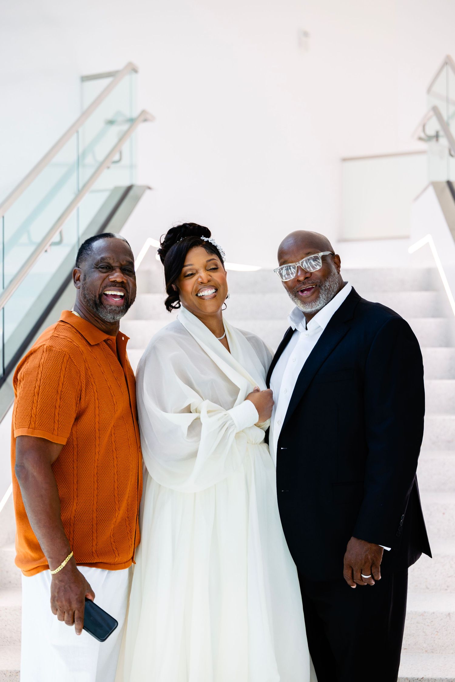 Joyful bride in a flowing white dress laughs between two smiling men, one in an orange shirt and one in a black suit, on a modern staircase.