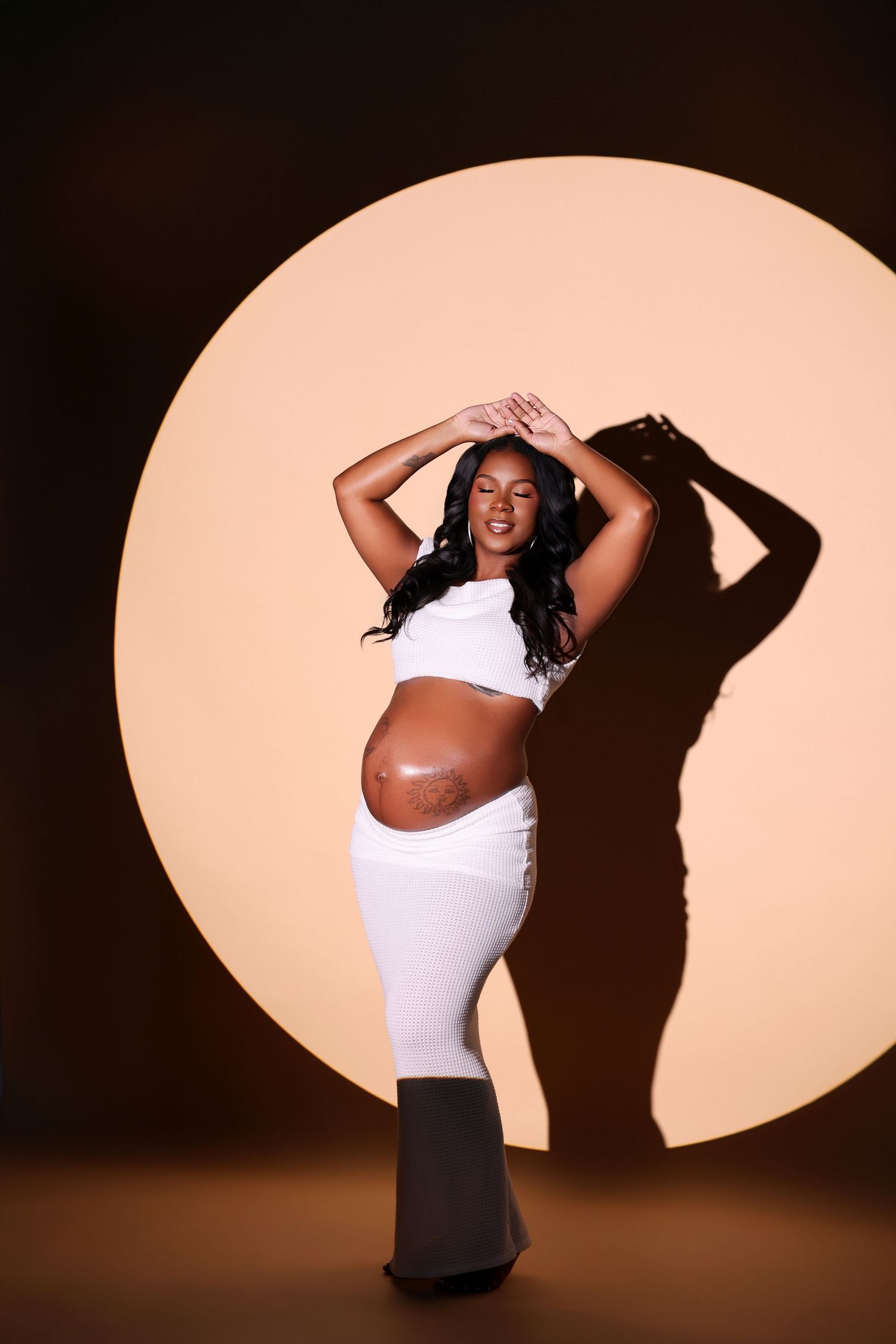 Pregnant woman in a white crop top and long skirt poses with arms raised in front of a peach spotlight circle backdrop.