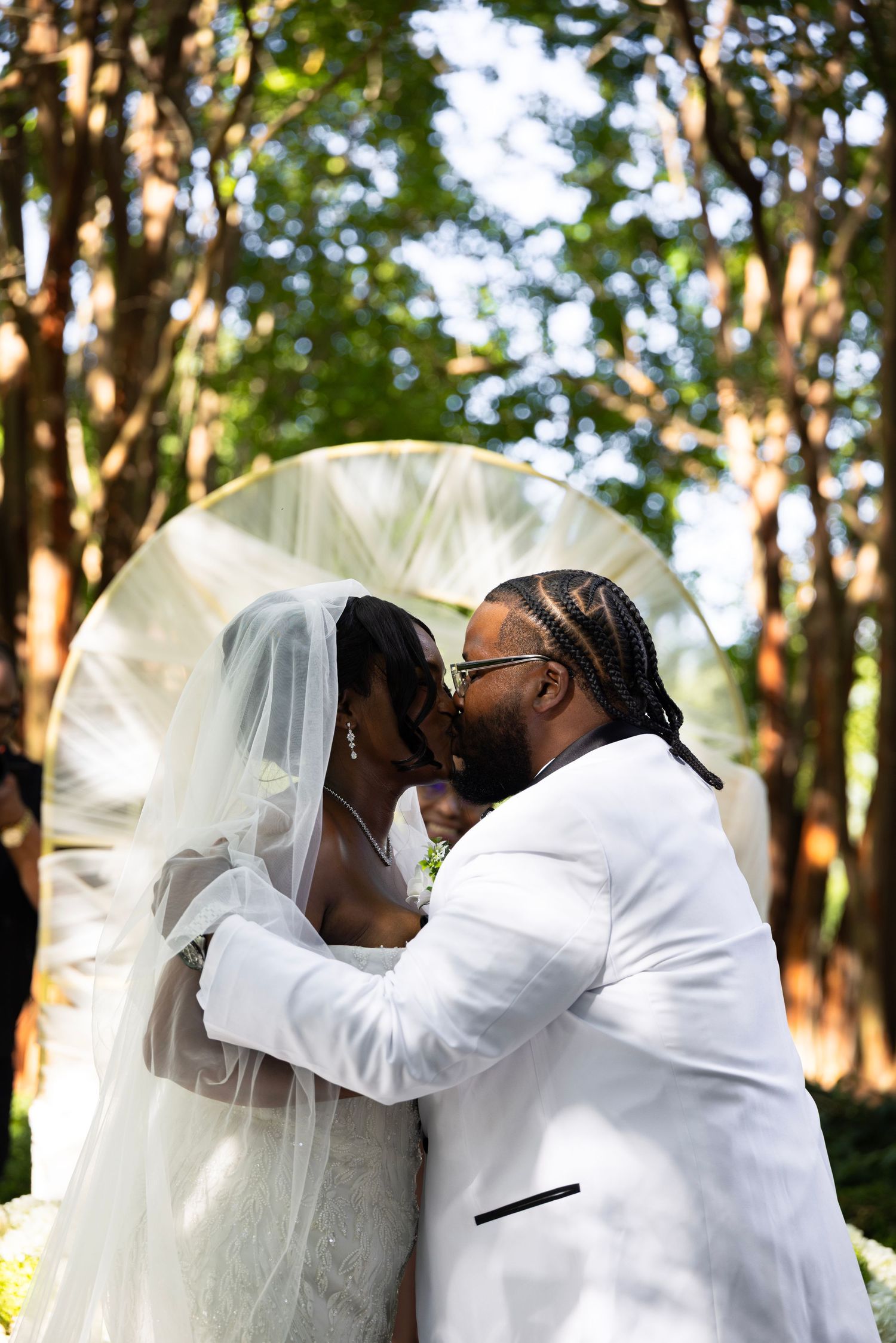 A bride in a long veil and a groom in a white tuxedo jacket share a kiss during an outdoor wedding ceremony beneath tall trees.