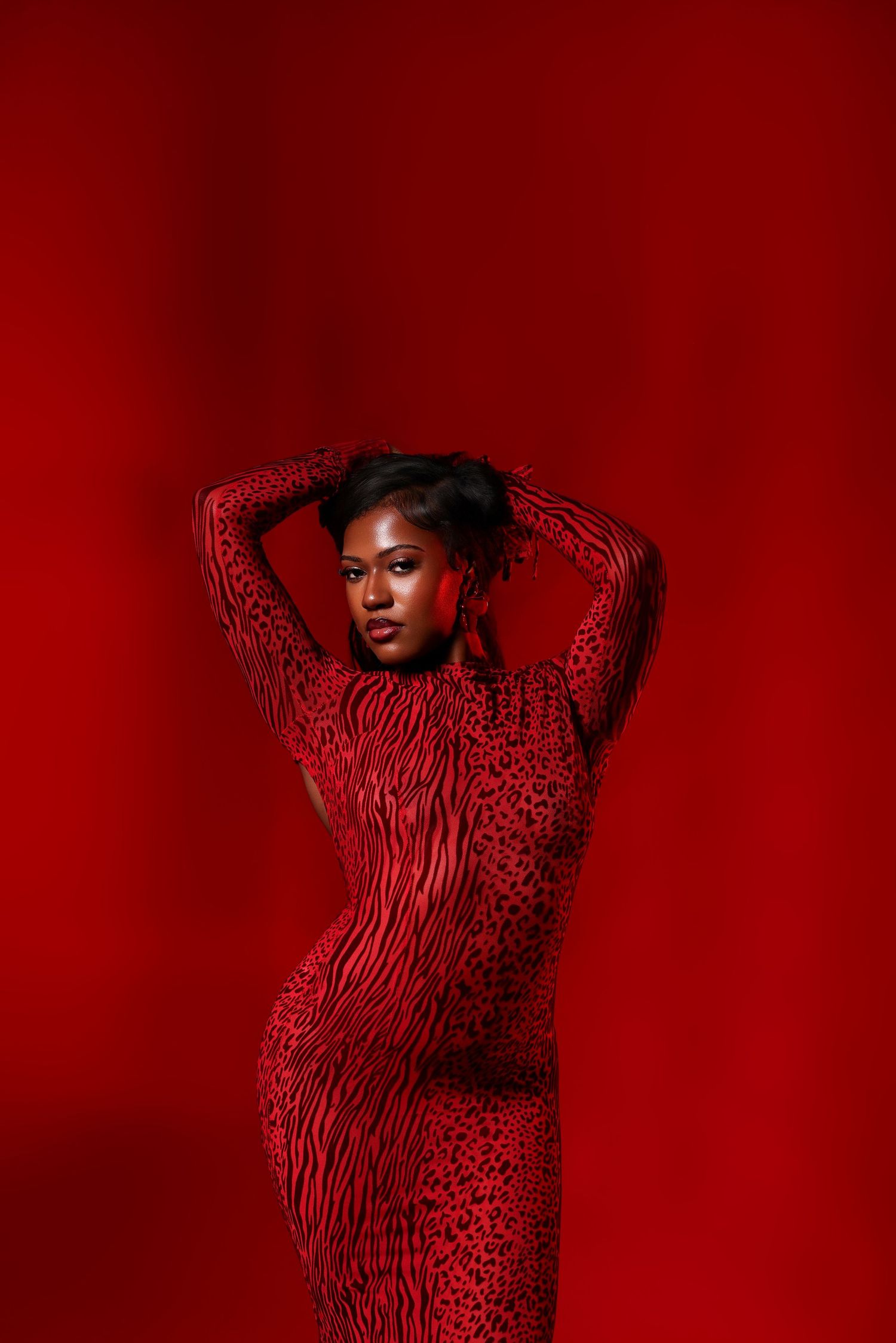 A woman in a red patterned bodycon dress poses with her arms raised overhead against a vivid red studio background.