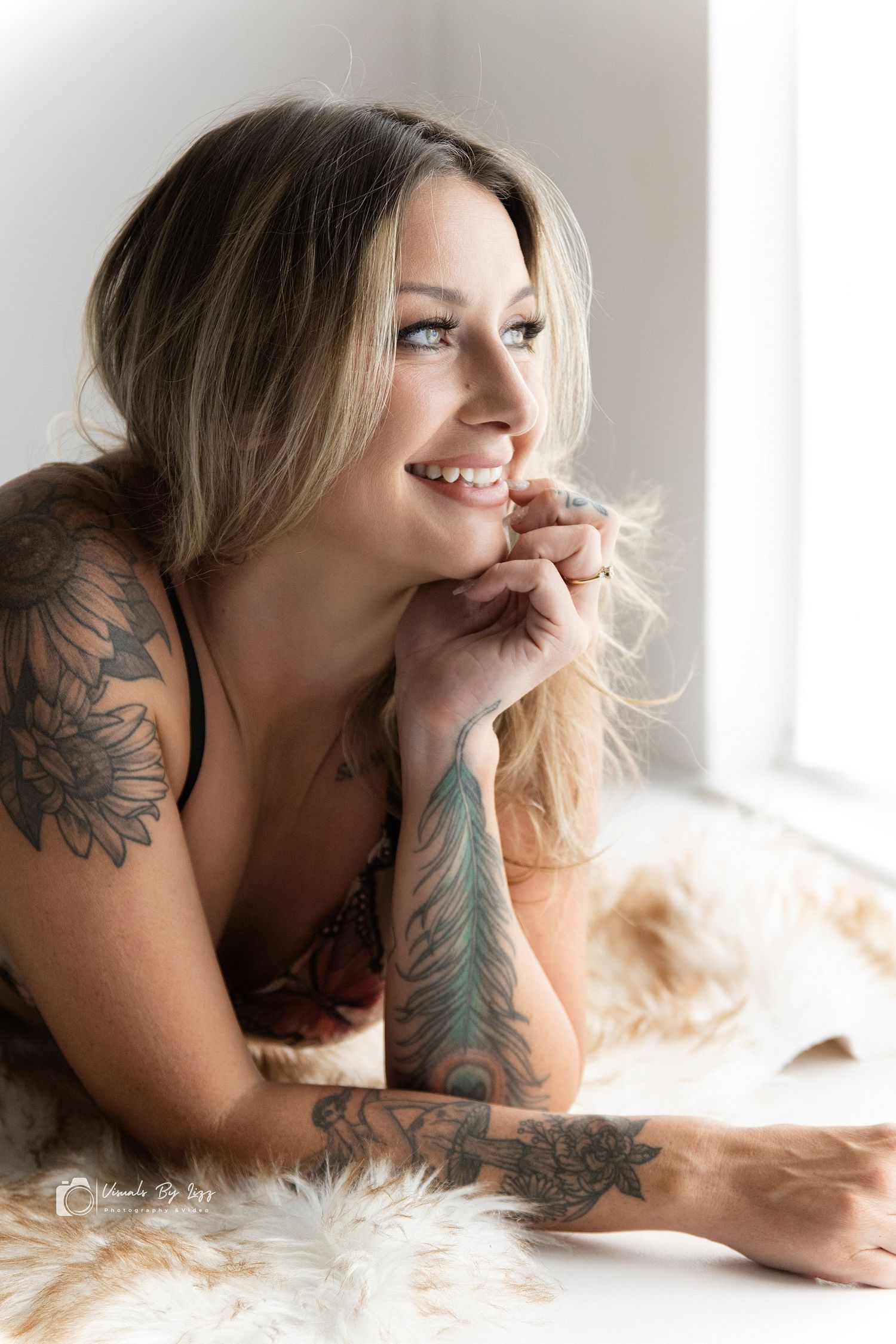Smiling blonde woman with floral tattoos on arms and shoulders poses near a bright window on a fluffy rug.
