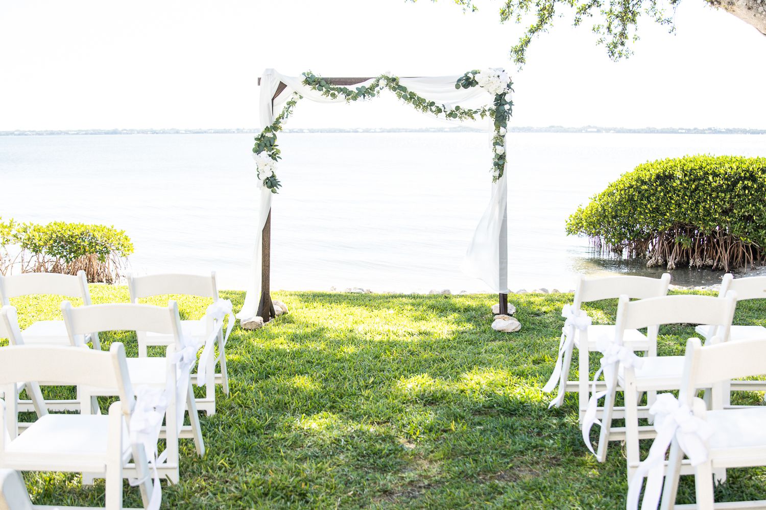 Outdoor wedding ceremony setup with white chairs and floral arch overlooking the ocean on a grassy lawn.
