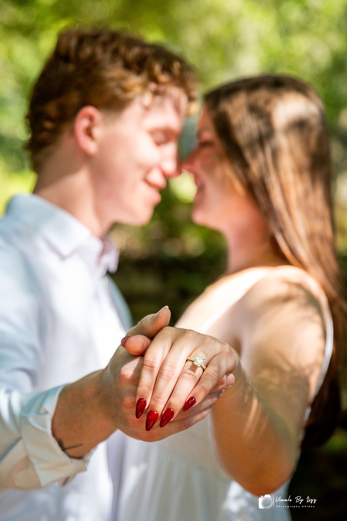 Couple sharing a romantic moment, showing off engagement ring with red nails, green bokeh background In Bok Tower Garden, Lake Wales, Florid