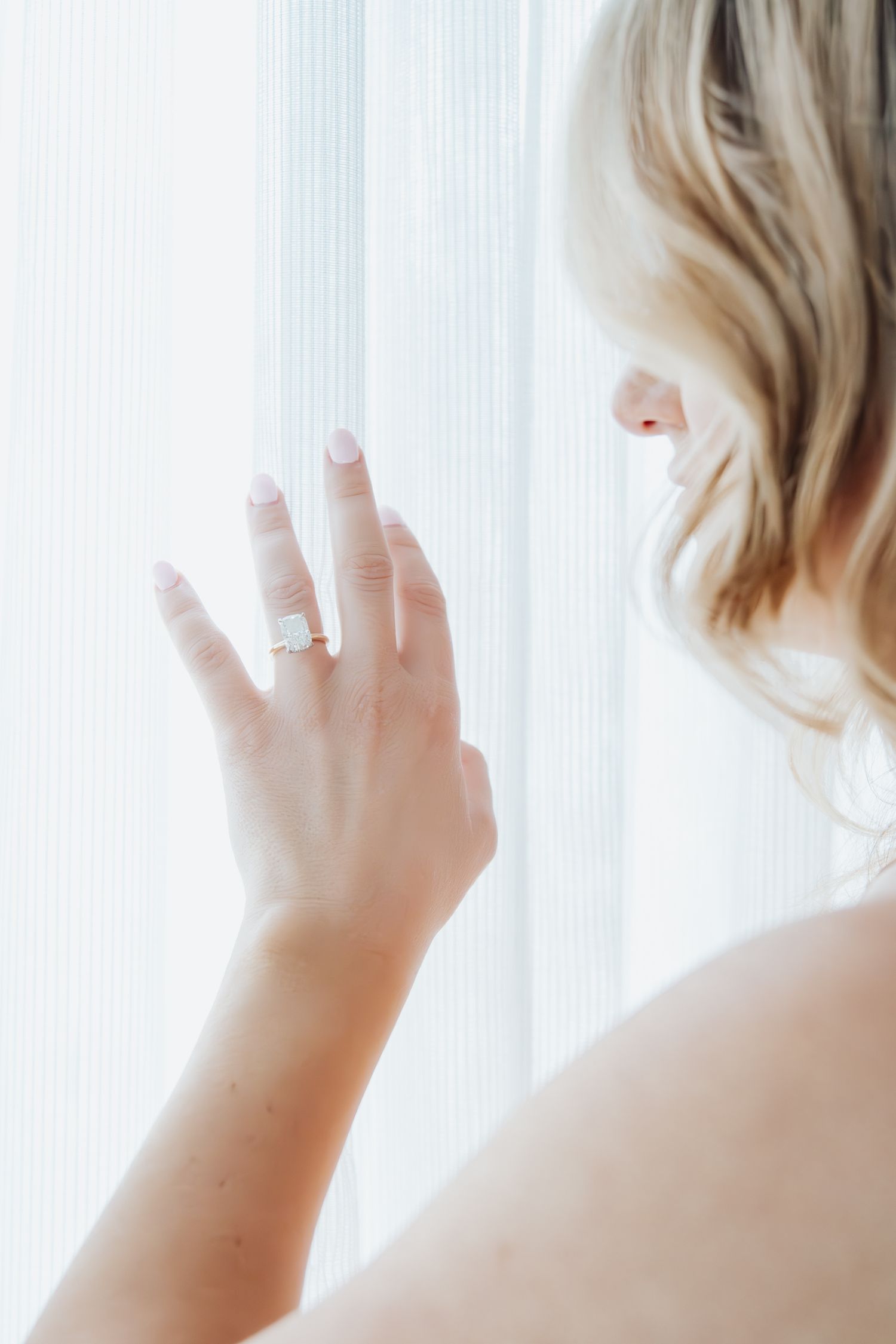 Soft backlit photo of engagement ring on hand against white curtains.