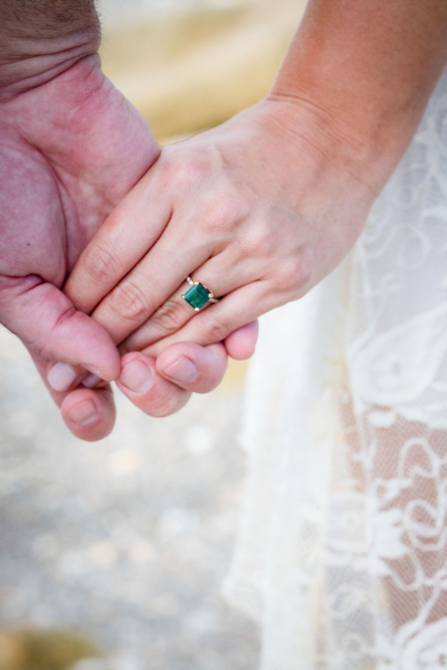 Close-up of hands holding together showing an emerald engagement ring with lace dress details.