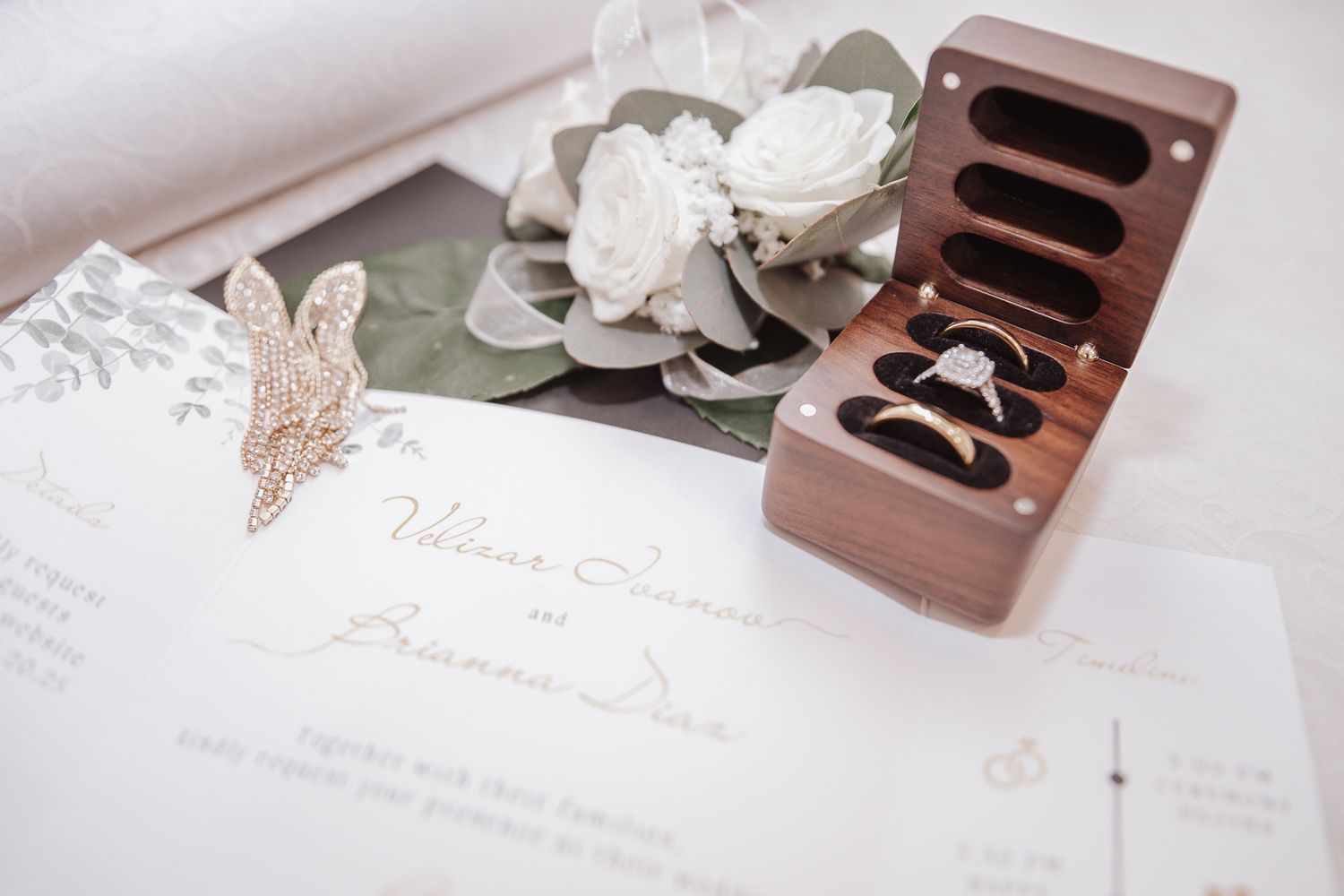 A rustic wooden ring box sits beside white floral decor and elegant wedding stationery on a light surface.