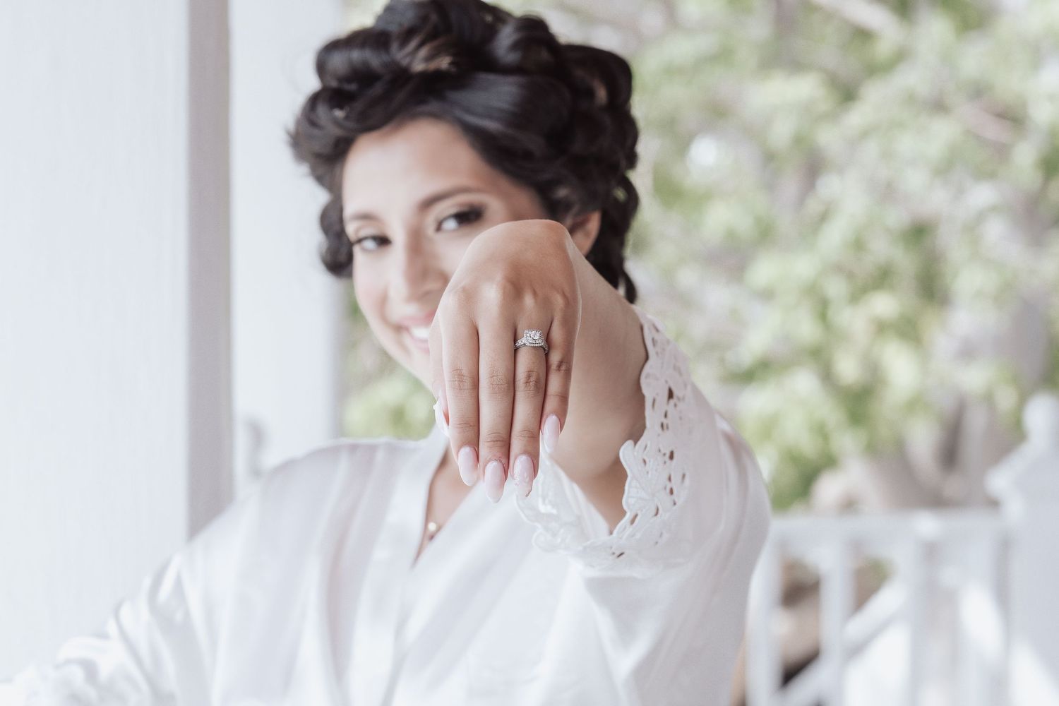 A person wearing a white silk robe shows off an engagement ring while touching their face.