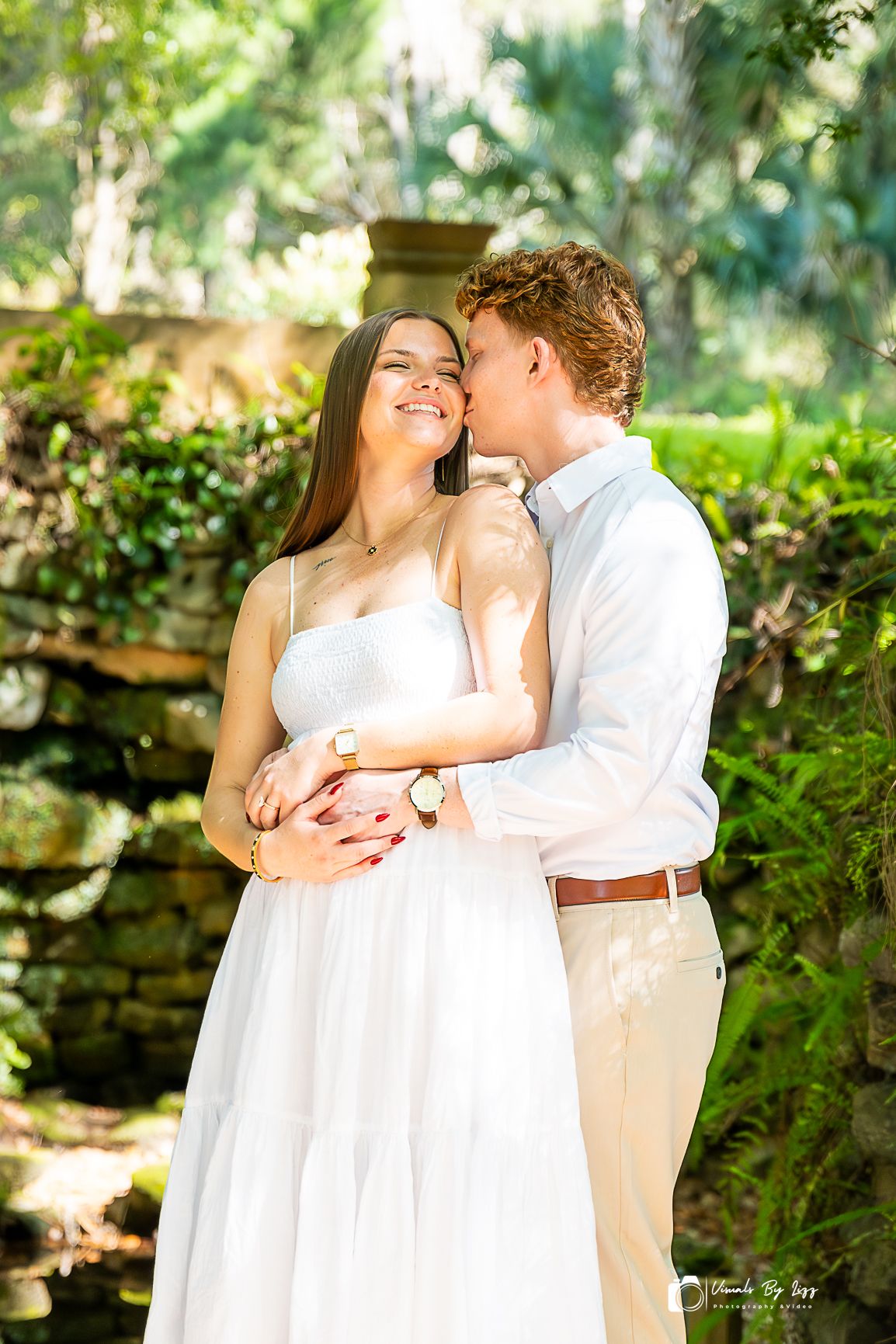 Couple embracing in a lush garden, woman in white dress smiling as man kisses her cheek during engagement photo session, Lake Wales, Florida