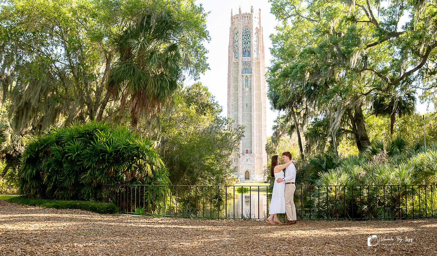 Couple embracing near ornate iron gates with Bok Tower rising above lush Spanish moss-draped trees in Lake Wales, Florida.