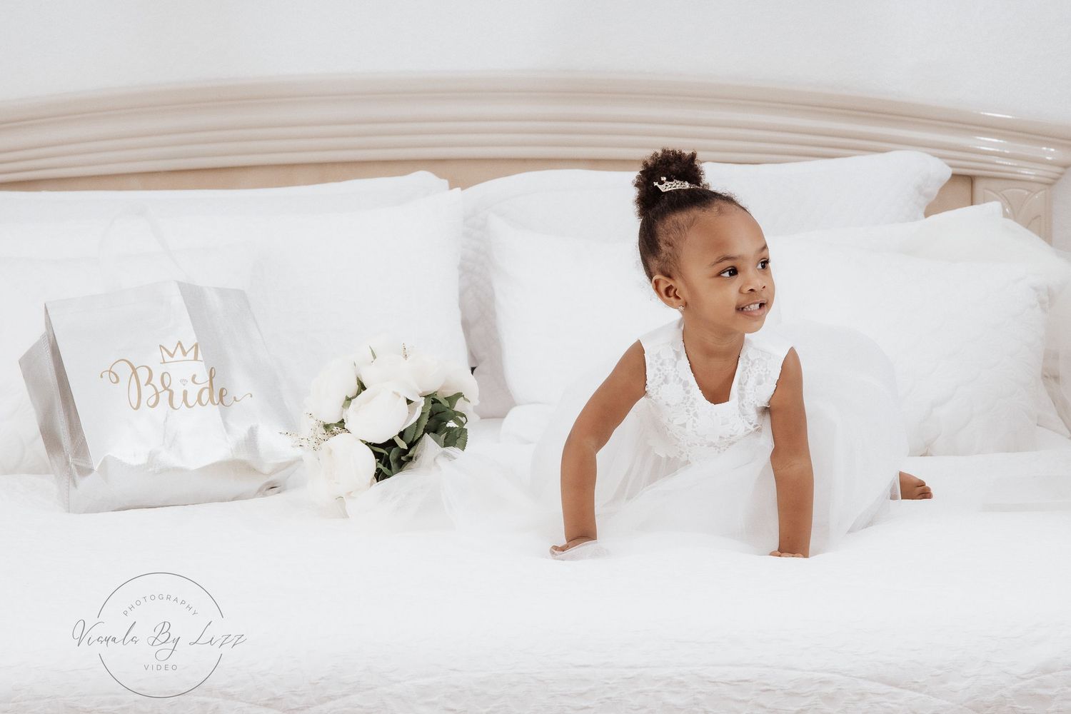 Small child in a white dress smile playfully while crawling on a white couch with decorative pillows and flowers.
