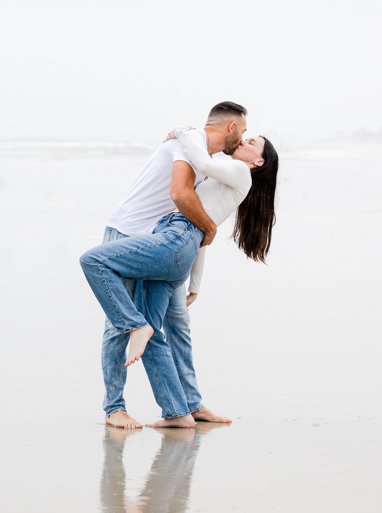 Couple sharing a romantic kiss on the beach, man lifting woman in jeans and white tops on wet sand.