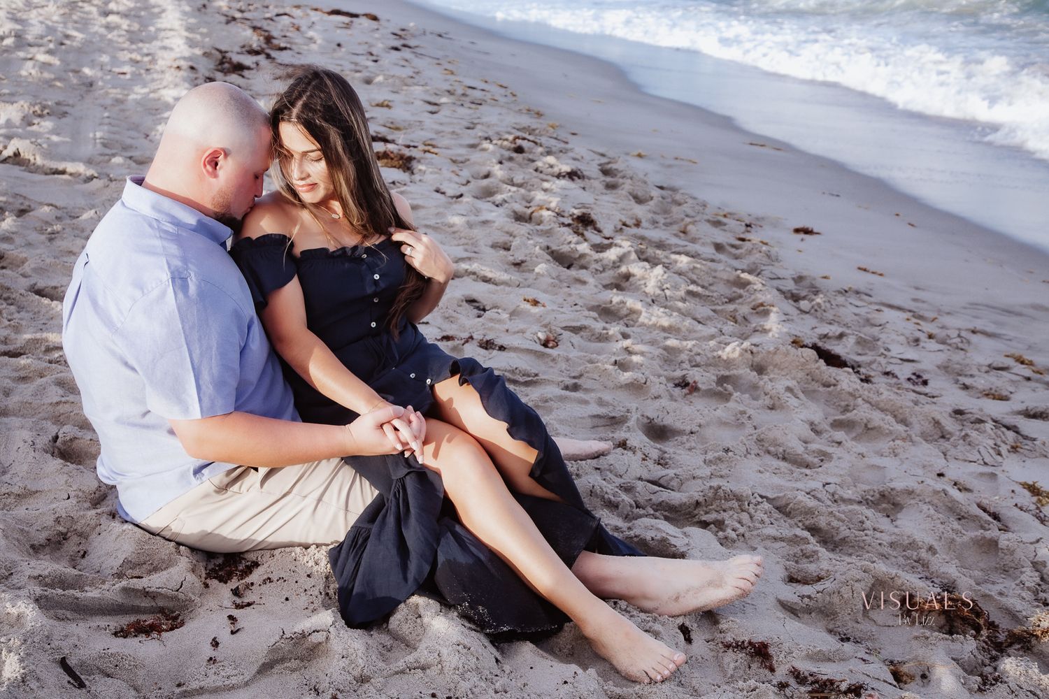 Couple sitting together on sandy beach near ocean waves, woman in black dress, man in blue shirt, romantic engagement photo Jupiter, FL