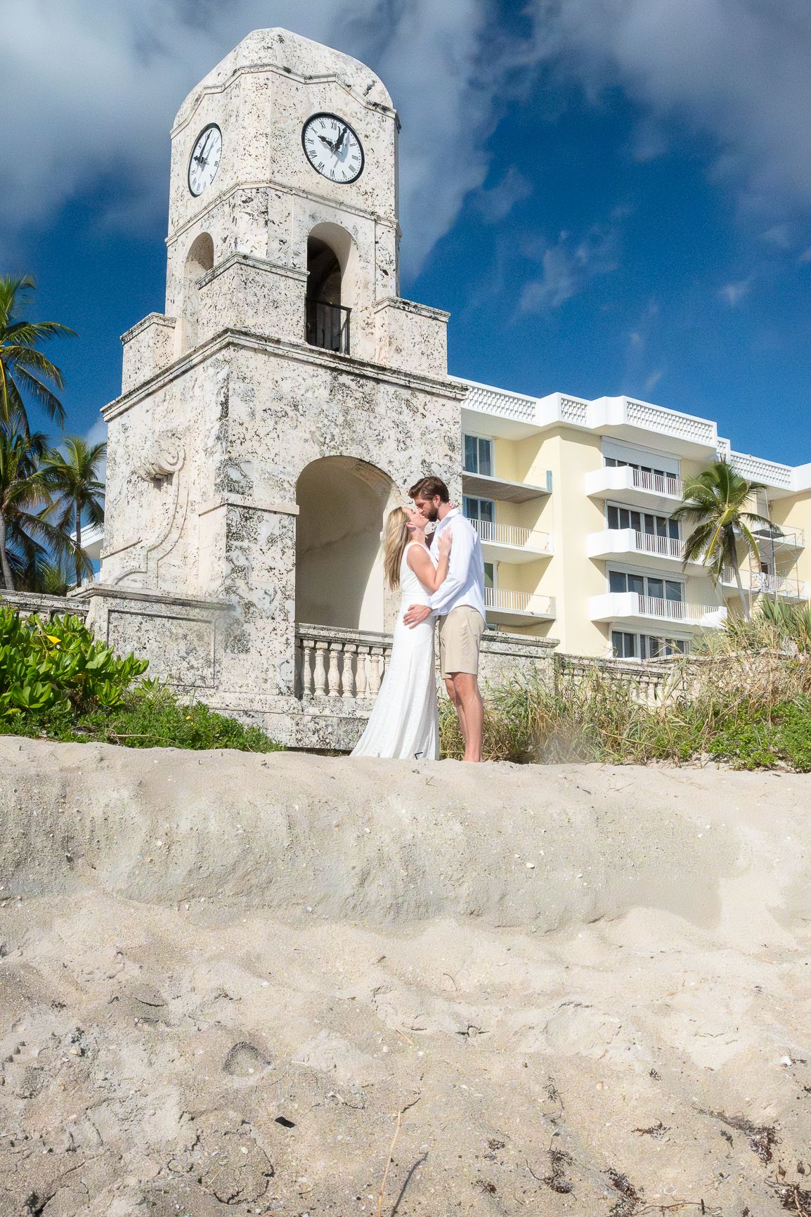 A romantic couple embraces near a white stone tower overlooking a tropical beach in Clock Tower, Worth Ave, Florida.