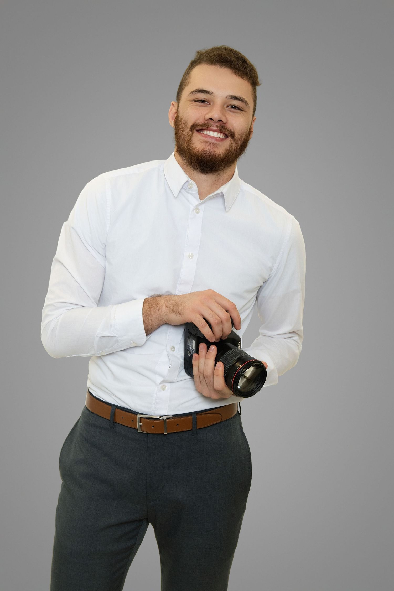 Professional photographer in white dress shirt and gray slacks holding camera equipment against studio backdrop.