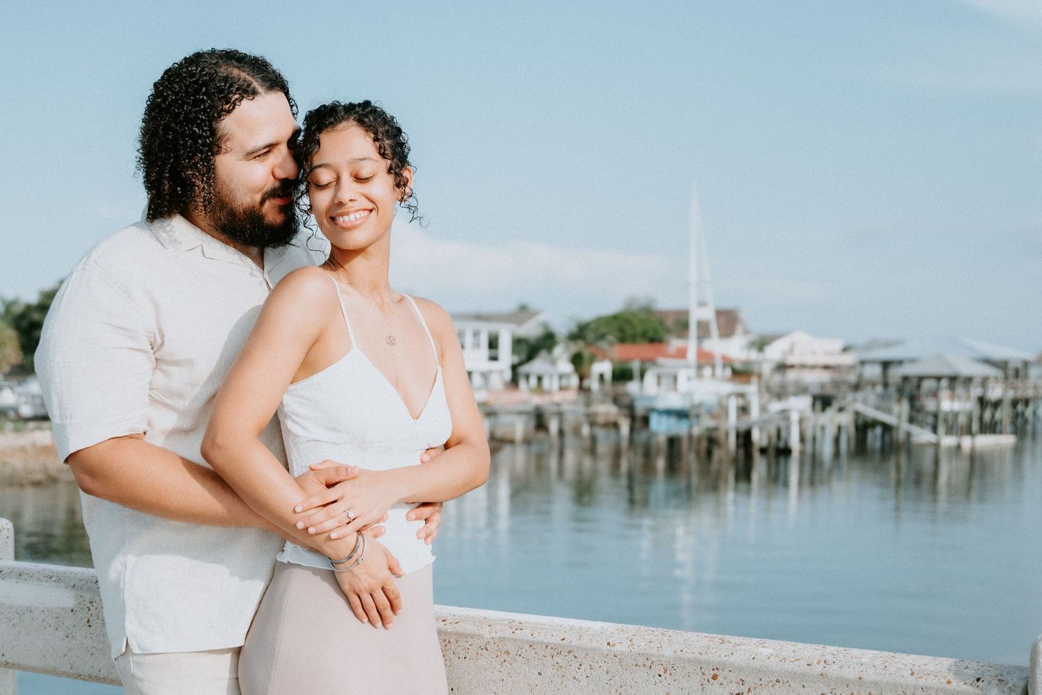 Couple in white outfits embracing near a waterfront marina on a sunny day.