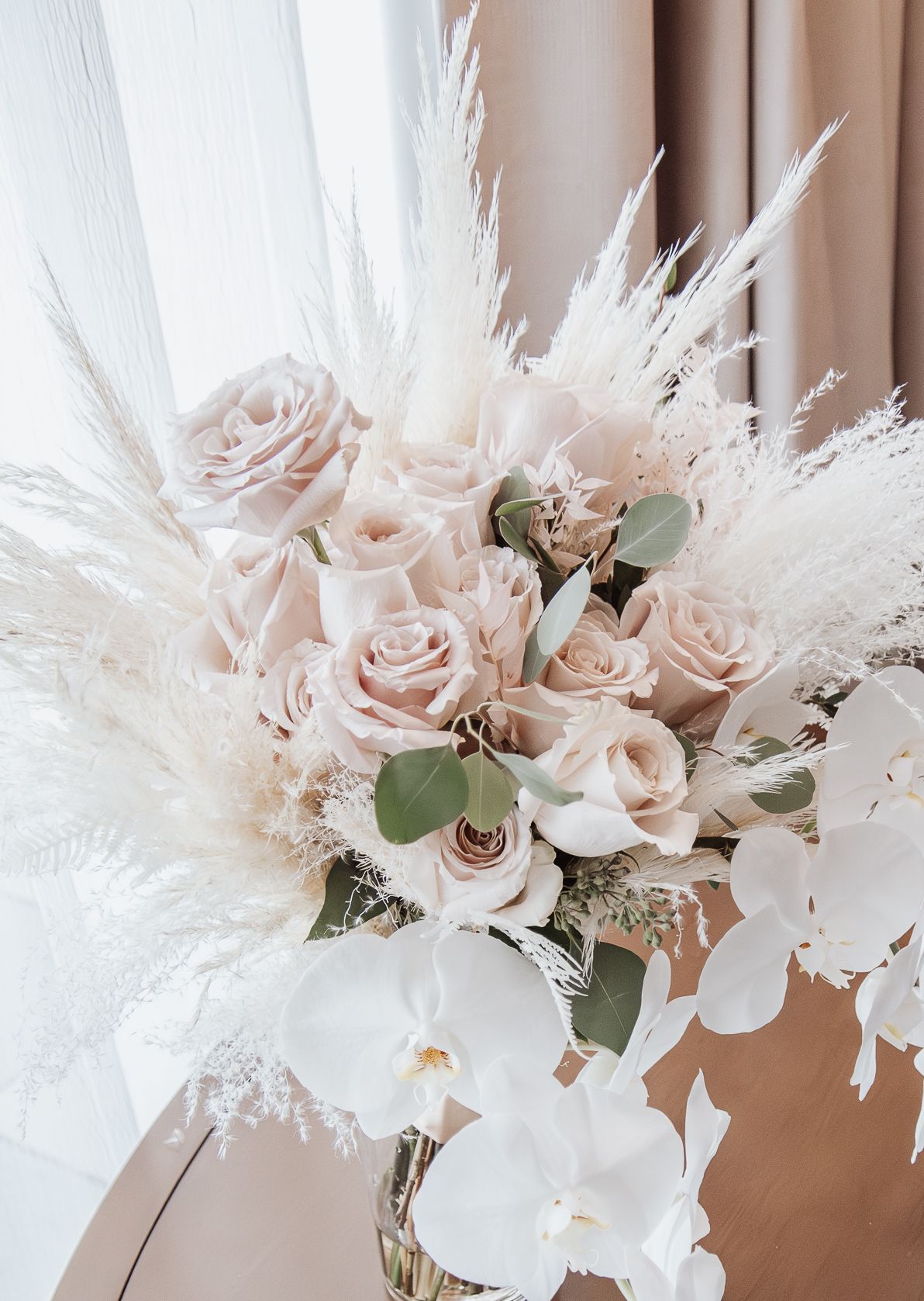 Elegant white and blush pink wedding bouquet with roses, pampas grass, and eucalyptus leaves against neutral backdrop.