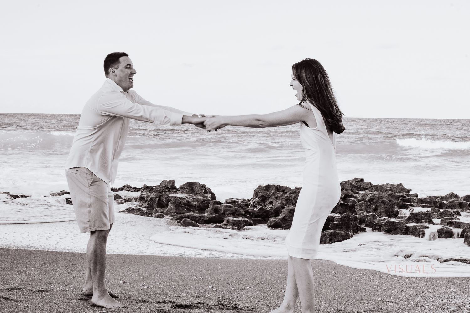 Romantic black and white beach photo of couple holding hands near rocky shore.
