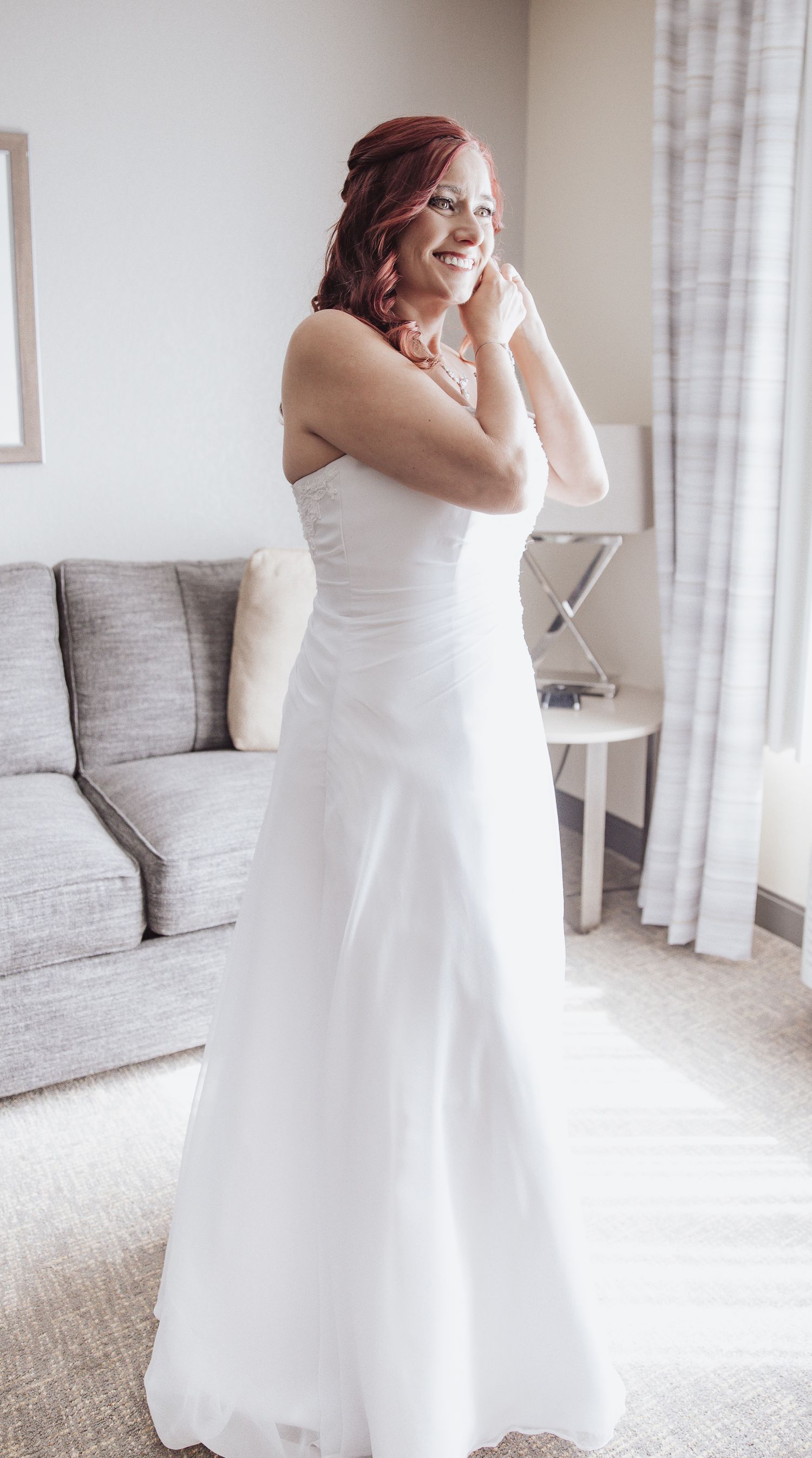 A white strapless wedding gown is worn with joy in a bright sunlit room with grey furniture.