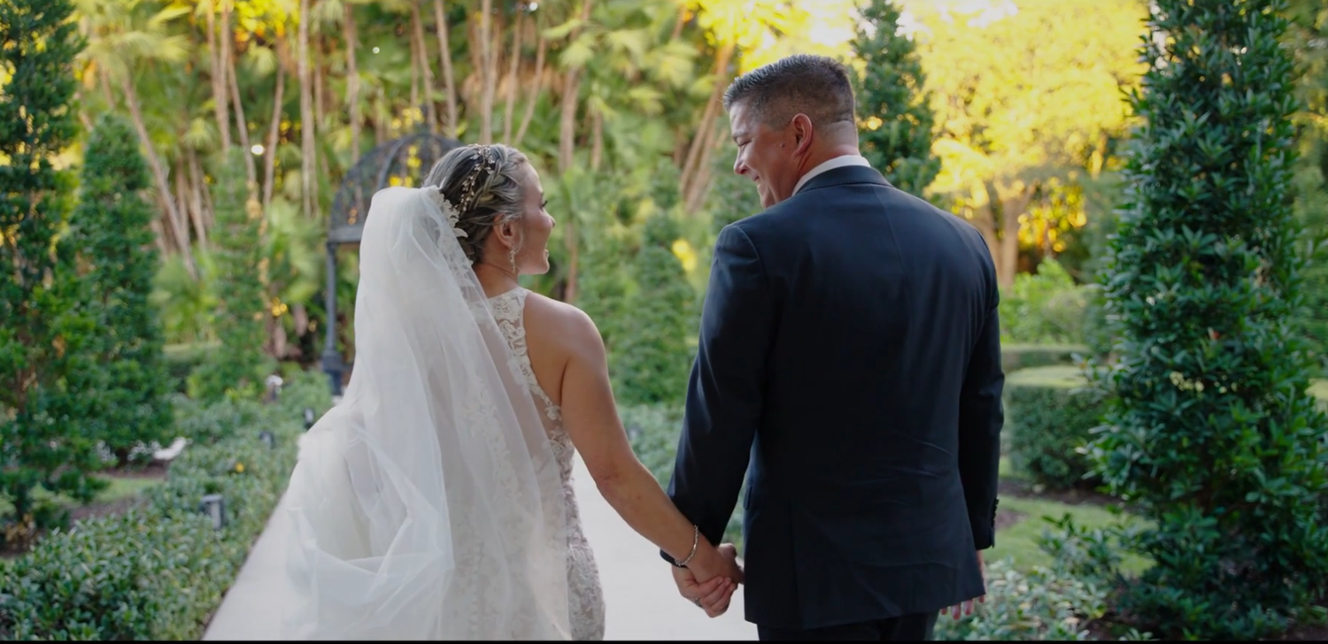 A couple in wedding attire walks hand in hand through a lush garden path surrounded by greenery and sunlight.