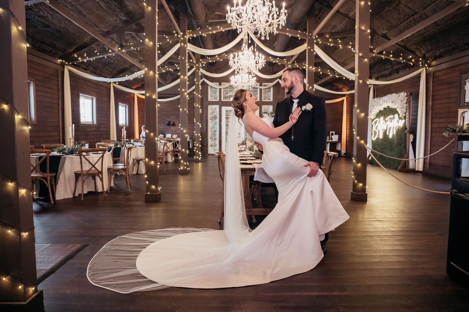 Elegant wedding couple dancing in a rustic barn venue decorated with twinkling lights and crystal chandeliers.